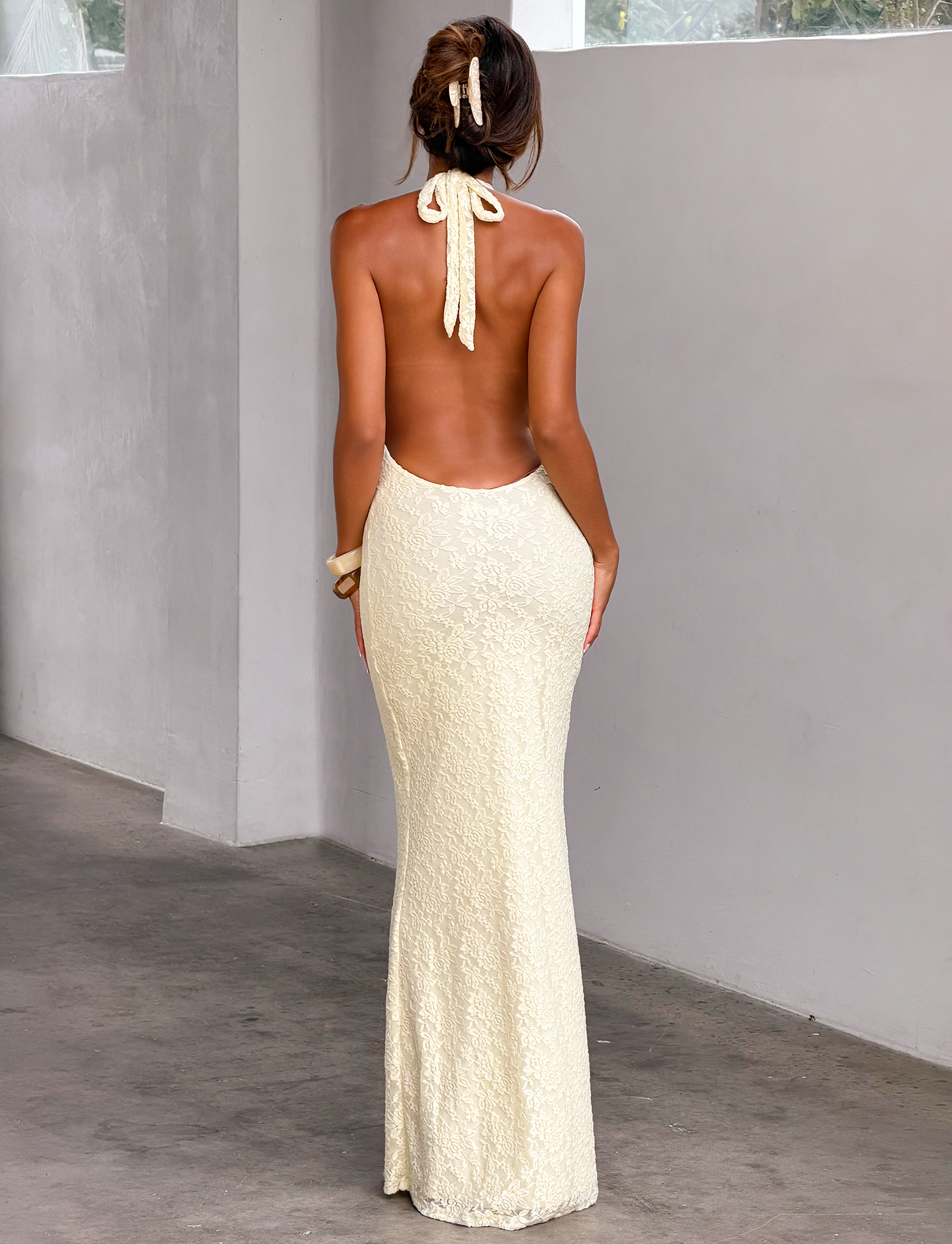 A woman, with her hair tied up in a matching ribbon, stands indoors near a gray wall and window wearing the TIGERMIST MAVEN LACE MAXI DRESS in pastel yellow. The dress features lace detailing, a halter neckline, and a bow at the neck.