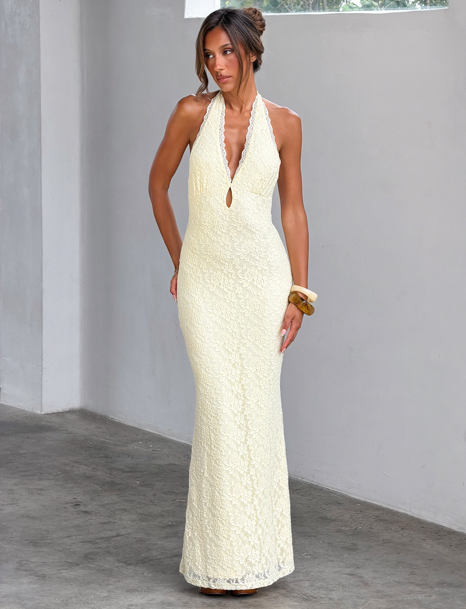 Indoors, a woman wears the TIGERMIST MAVEN LACE MAXI DRESS in pastel yellow, featuring a deep V-neckline and keyhole cutout. Her hair is styled up and one arm rests at her side.