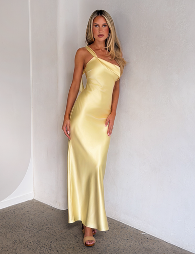 A woman with long blonde hair stands against a white wall, wearing the TIGERMIST RAINEY MAXI DRESS in butter yellow—a shiny, one-shoulder satin gown paired with open-toe heels.