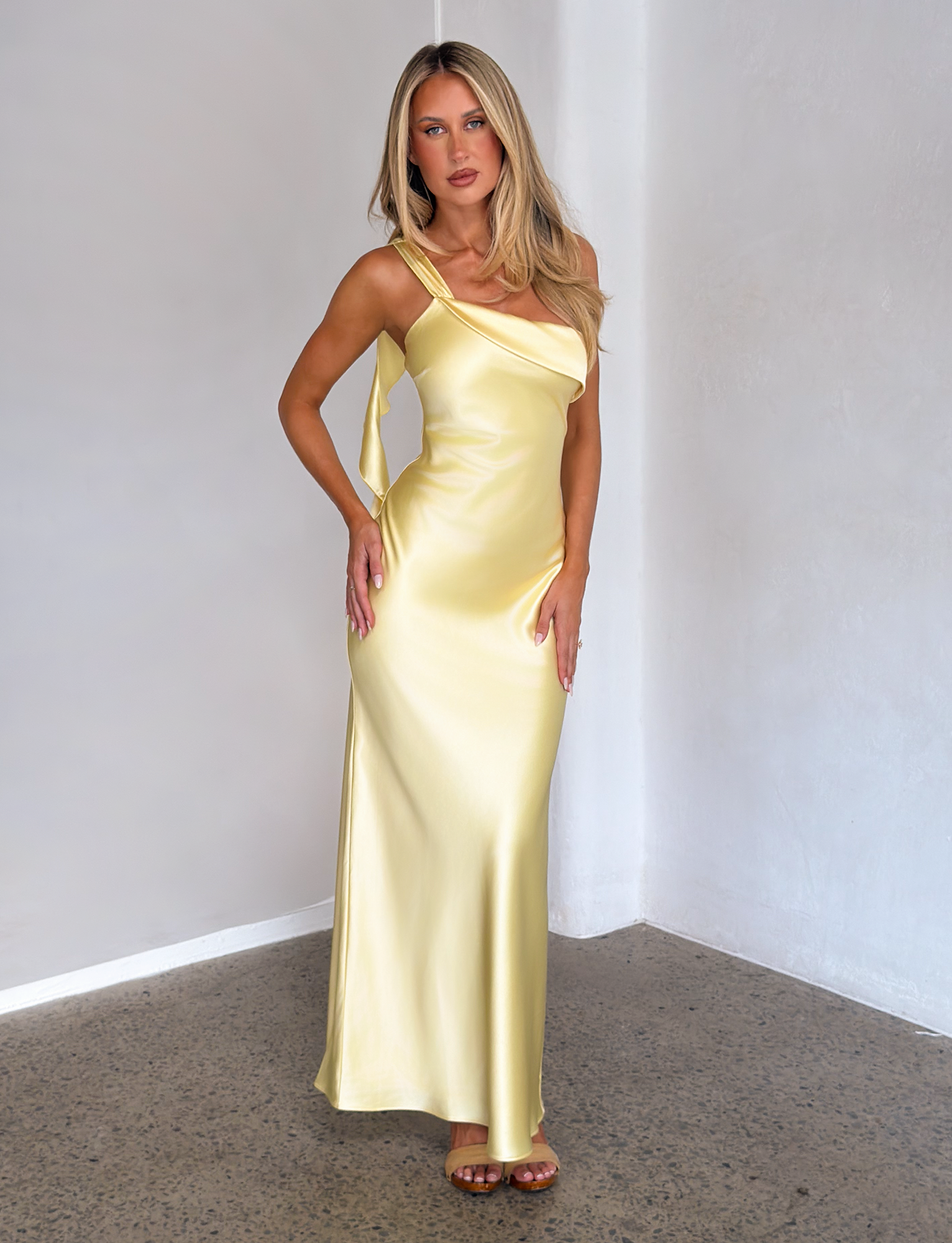 A woman with long blonde hair wears the TIGERMIST RAINEY MAXI DRESS in butter yellow, styled with brown sandals, standing indoors against a plain white wall and concrete floor.