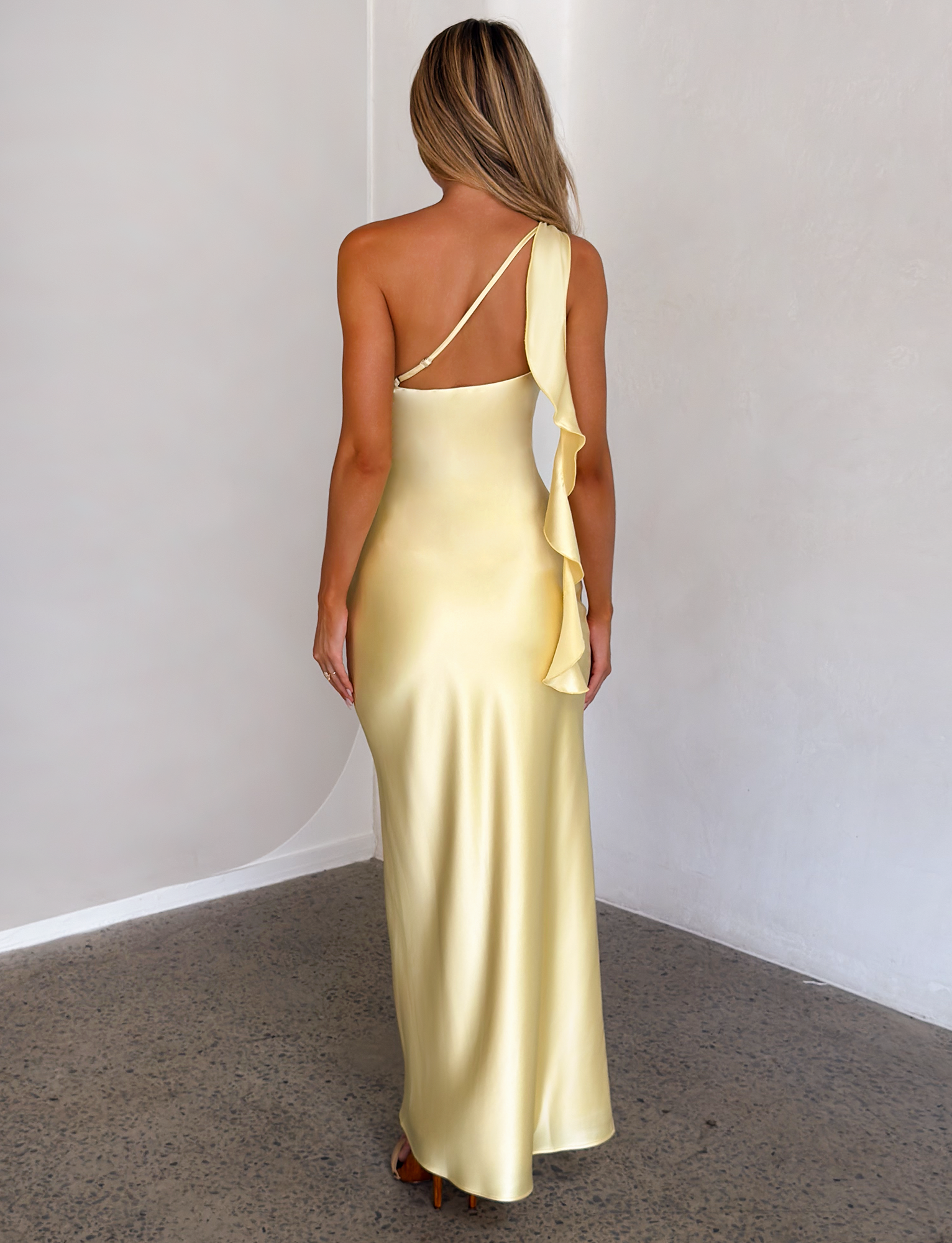 A woman in the TIGERMIST RAINEY MAXI DRESS in butter yellow, featuring a one-shoulder satin design with a waterfall ruffle, stands facing away in a minimalist room with light walls and a speckled floor.