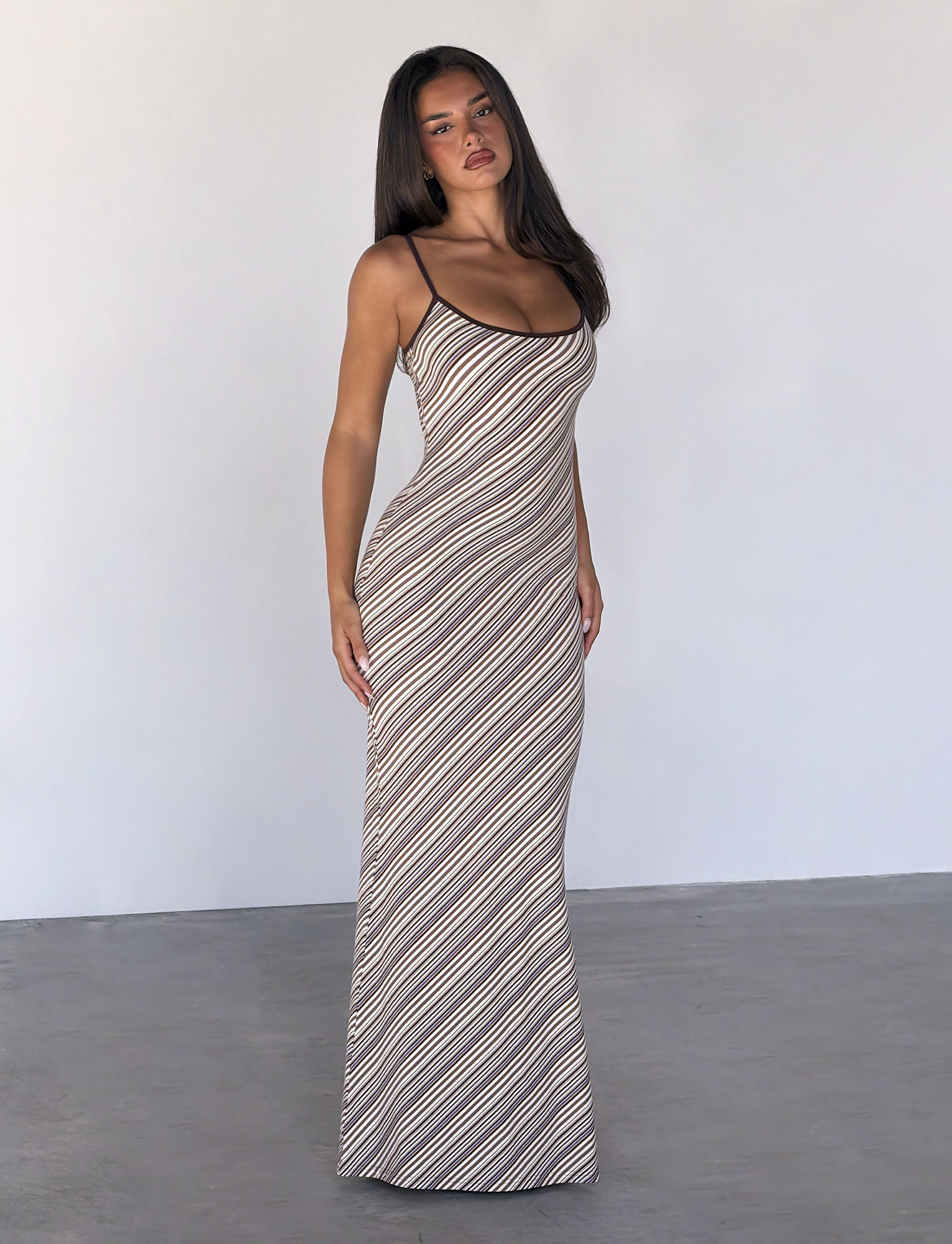 A woman with long dark hair and a neutral expression stands indoors against a plain light gray wall, wearing the TIGERMIST SLOANE MAXI DRESS in Tan and Choc Stripe with adjustable spaghetti straps.