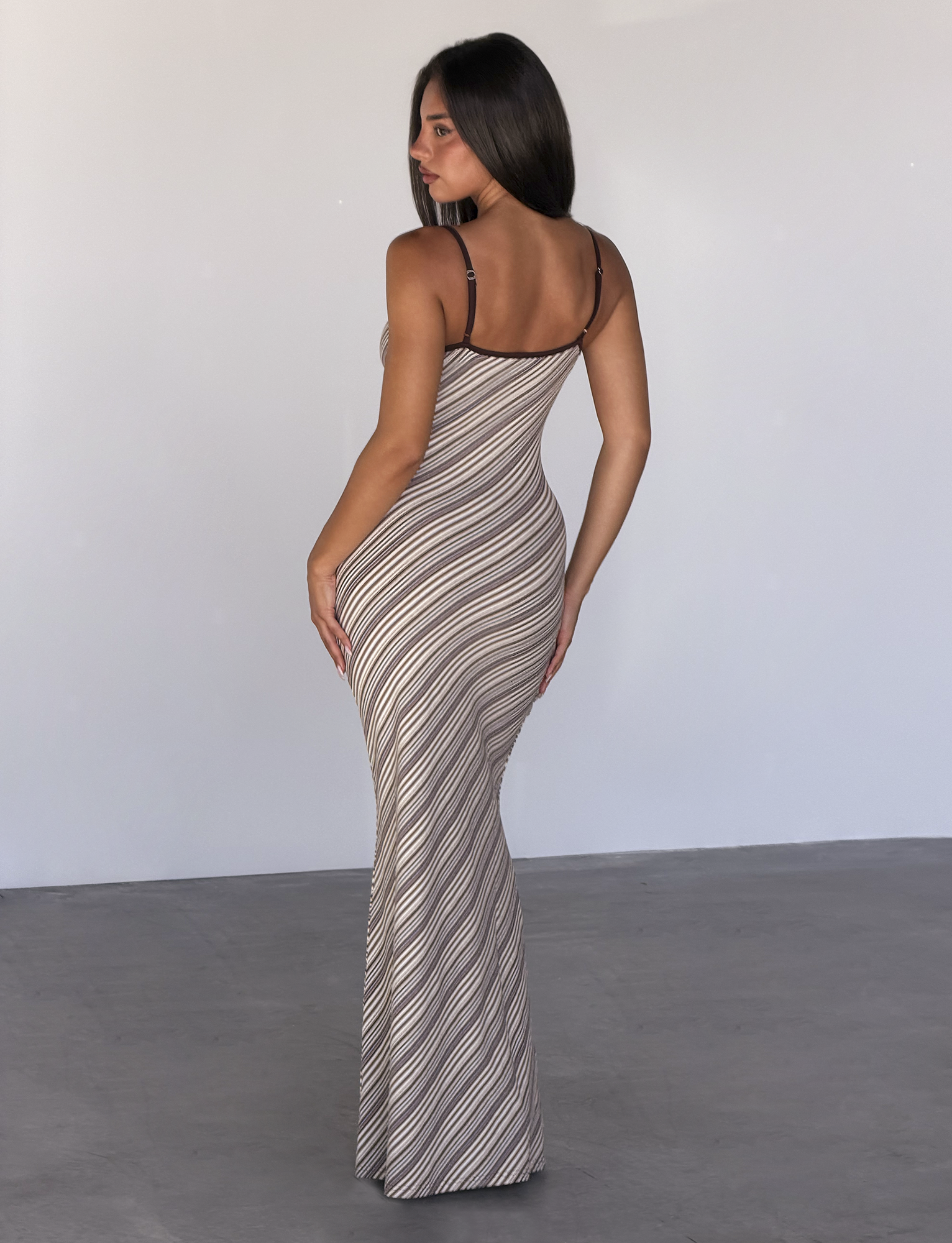A woman with long brown hair faces away, wearing the TIGERMIST SLOANE MAXI DRESS in Tan and Choc Stripe—fitted and sleeveless with adjustable spaghetti straps—standing on a smooth gray floor against a light-colored wall.