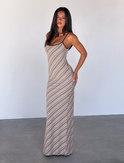 A woman poses indoors against a light wall, wearing the TIGERMIST SLOANE MAXI DRESS - TAN AND CHOC STRIPE. The sleeveless, figure-hugging dress features adjustable spaghetti straps and diagonal stripes, accentuating her silhouette.