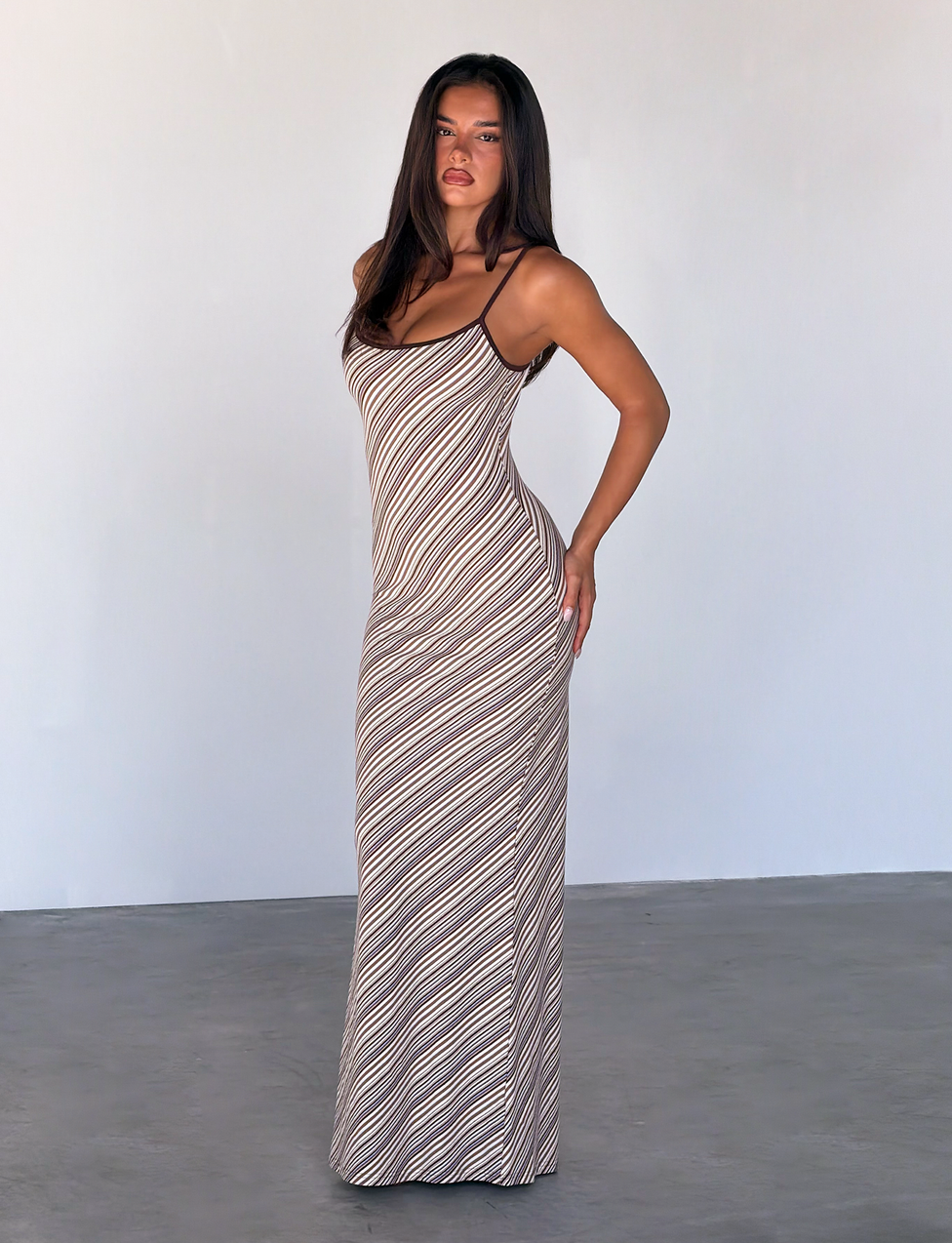 A woman poses indoors against a light wall, wearing the TIGERMIST SLOANE MAXI DRESS - TAN AND CHOC STRIPE. The sleeveless, figure-hugging dress features adjustable spaghetti straps and diagonal stripes, accentuating her silhouette.