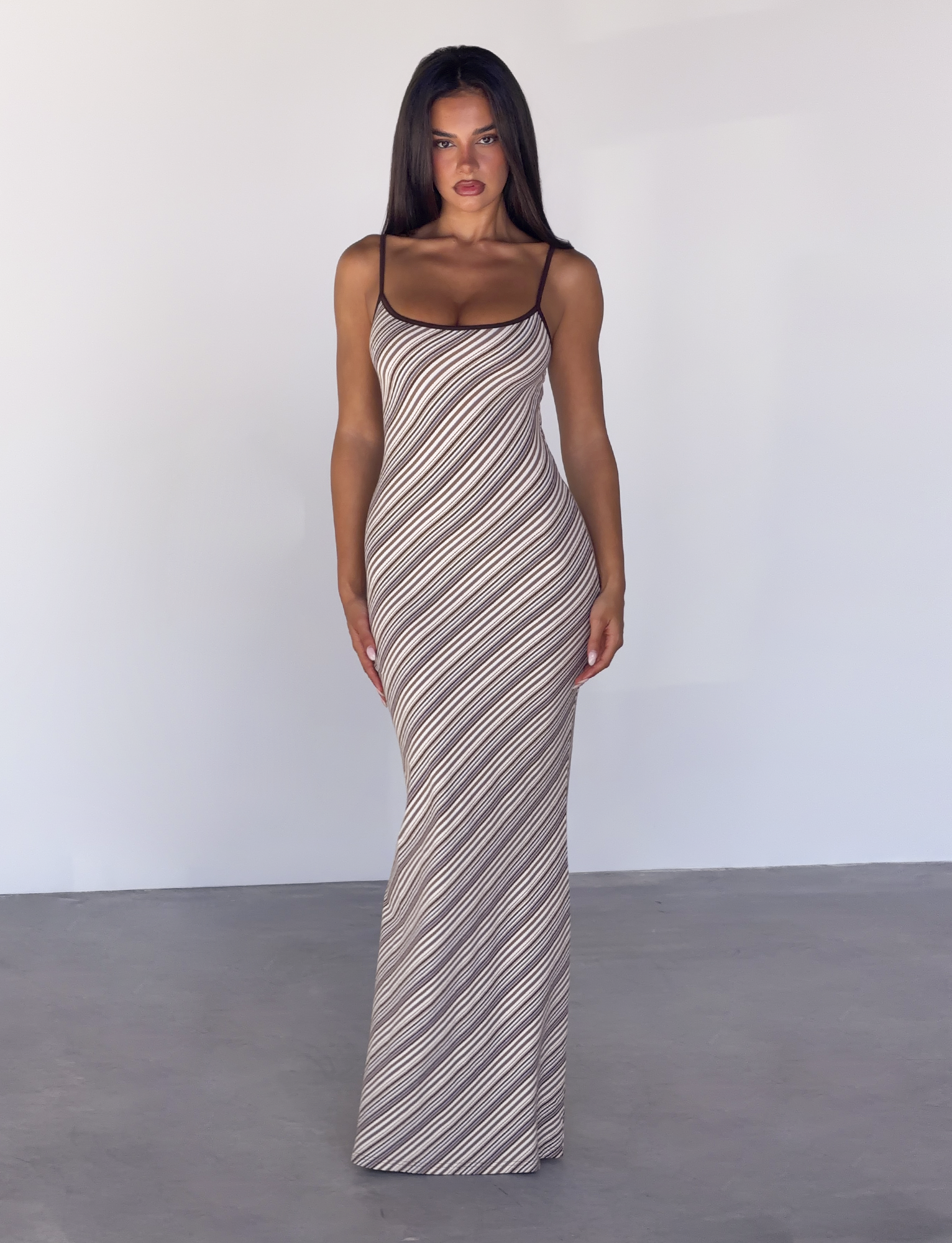 A woman with straight brown hair wears the TIGERMIST SLOANE MAXI DRESS - TAN AND CHOC STRIPE, a fitted, floor-length dress with diagonal stripes and adjustable spaghetti straps, standing against a white wall and gray floor.