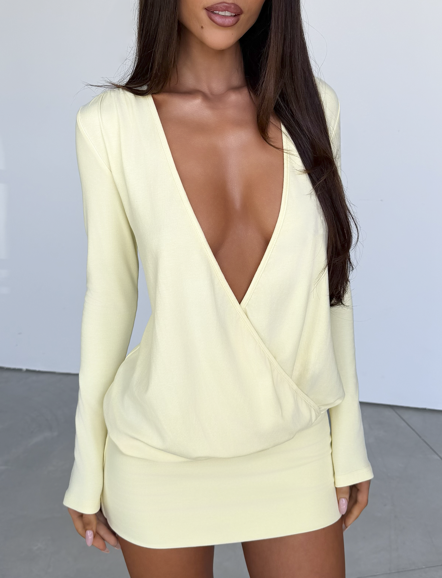 A woman stands against a plain background wearing the TIGERMIST FOXLEY MINI DRESS in Butter Cream—a pale yellow, long-sleeved mini dress featuring a deep V neckline and elegant wrap surplice silhouette. Her face is partially out of frame as the dress drapes beautifully on her figure.