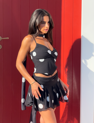 A woman with long dark hair wears the TIGERMIST AYDIN MINI SKIRT - BLACK and a black sleeveless crop top, standing in sunlight before a red door with a white section on the right.