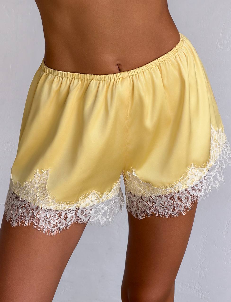A person wearing the SOLSTICE SHORT- YELLOW by TIGERMIST, featuring yellow satin fabric and white lace trim at the hem, stands against a light-colored background.