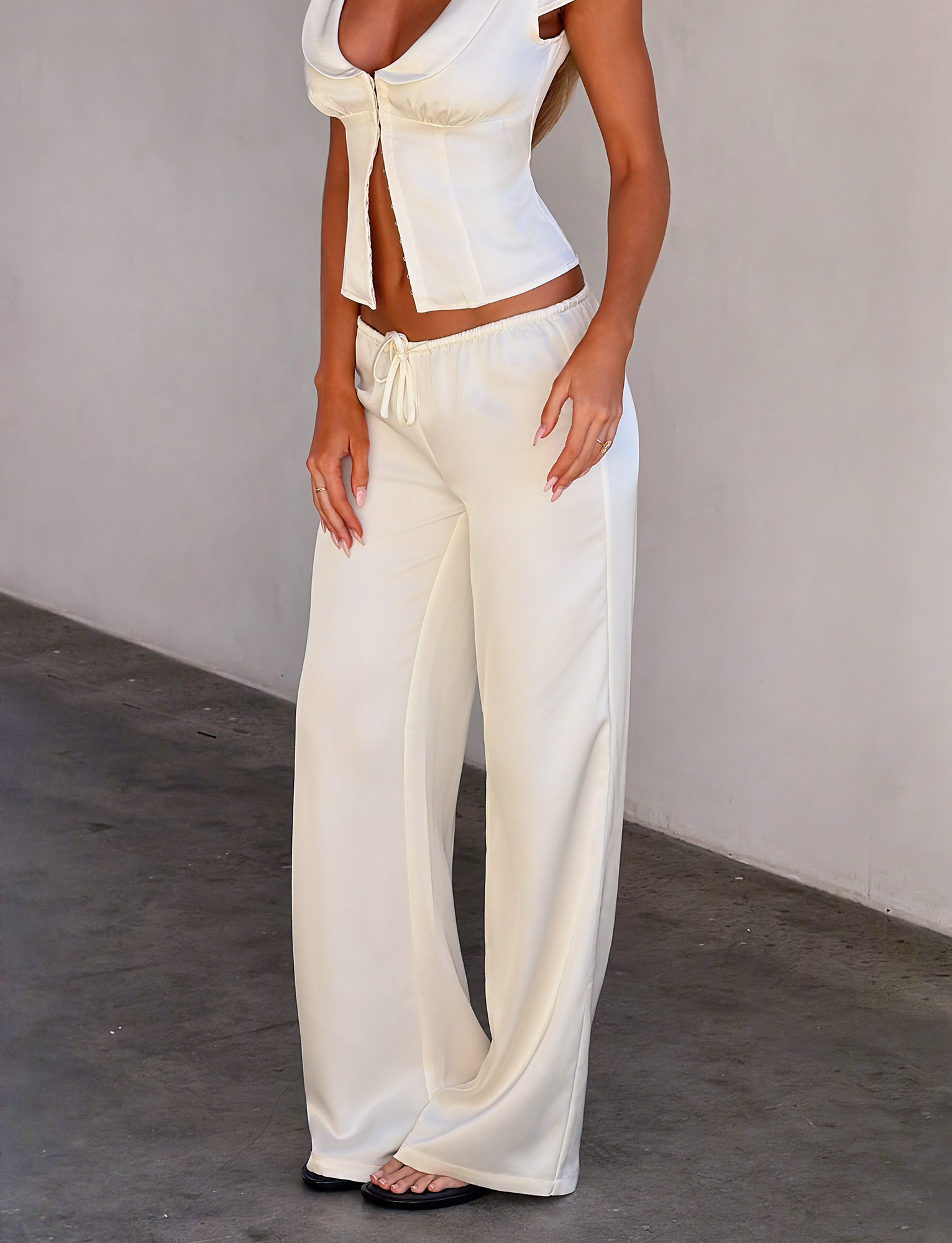 A woman stands against a plain wall with her hands by her sides, wearing the TIGERMIST MAURA PANT in white. She pairs the loose, wide-legged pants with a sleeveless white crop top featuring tie details. Her face is not visible in the image.