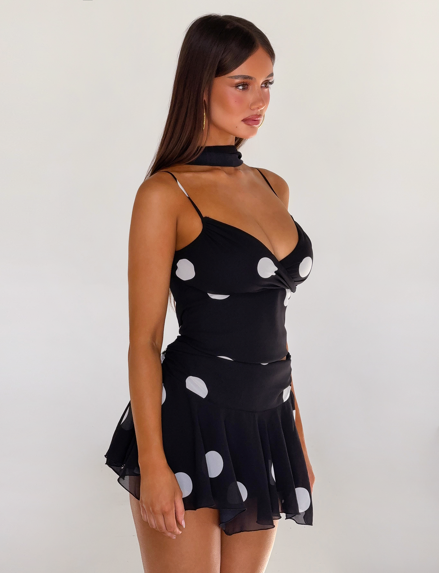 A woman with long brown hair models the TIGERMIST AYDIN SCARF TOP - BLACK, featuring a black chiffon triangle cami with white polka dots and adjustable straps, paired with a matching neck scarf against a plain light background.