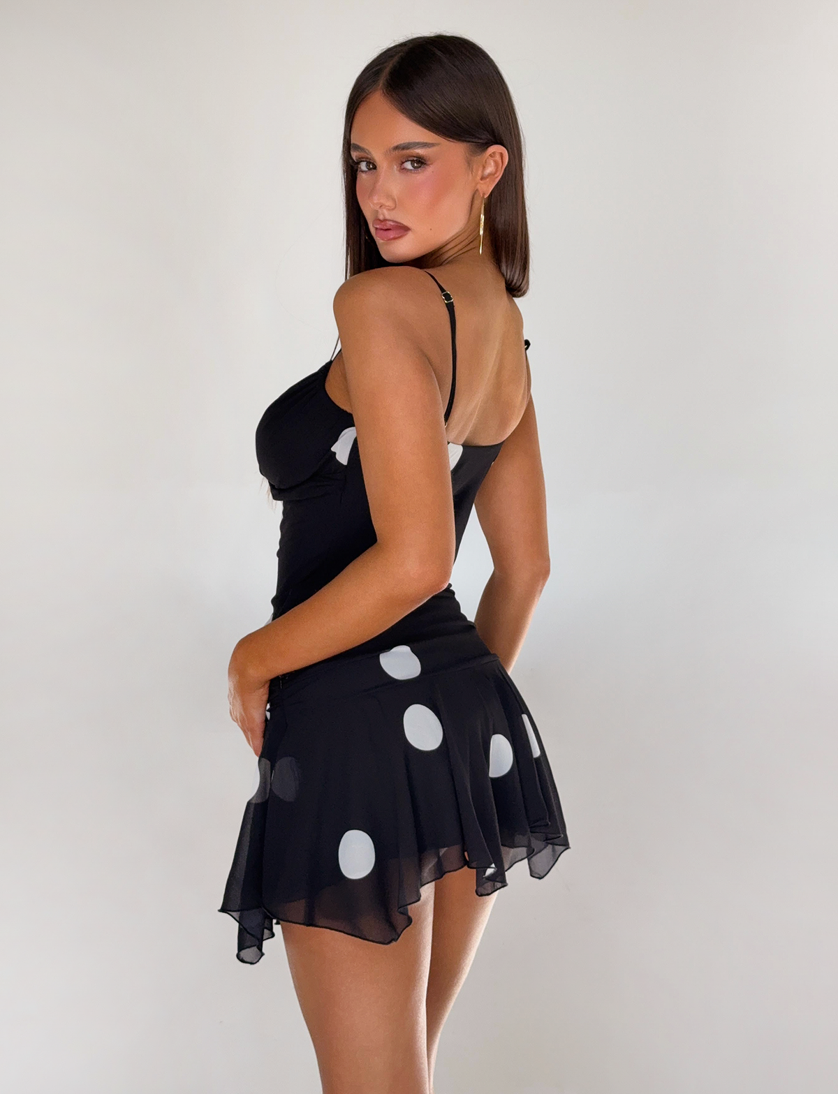 A woman with long brown hair models the TIGERMIST AYDIN MINI SKIRT - BLACK, featuring thin straps, against a plain white background.