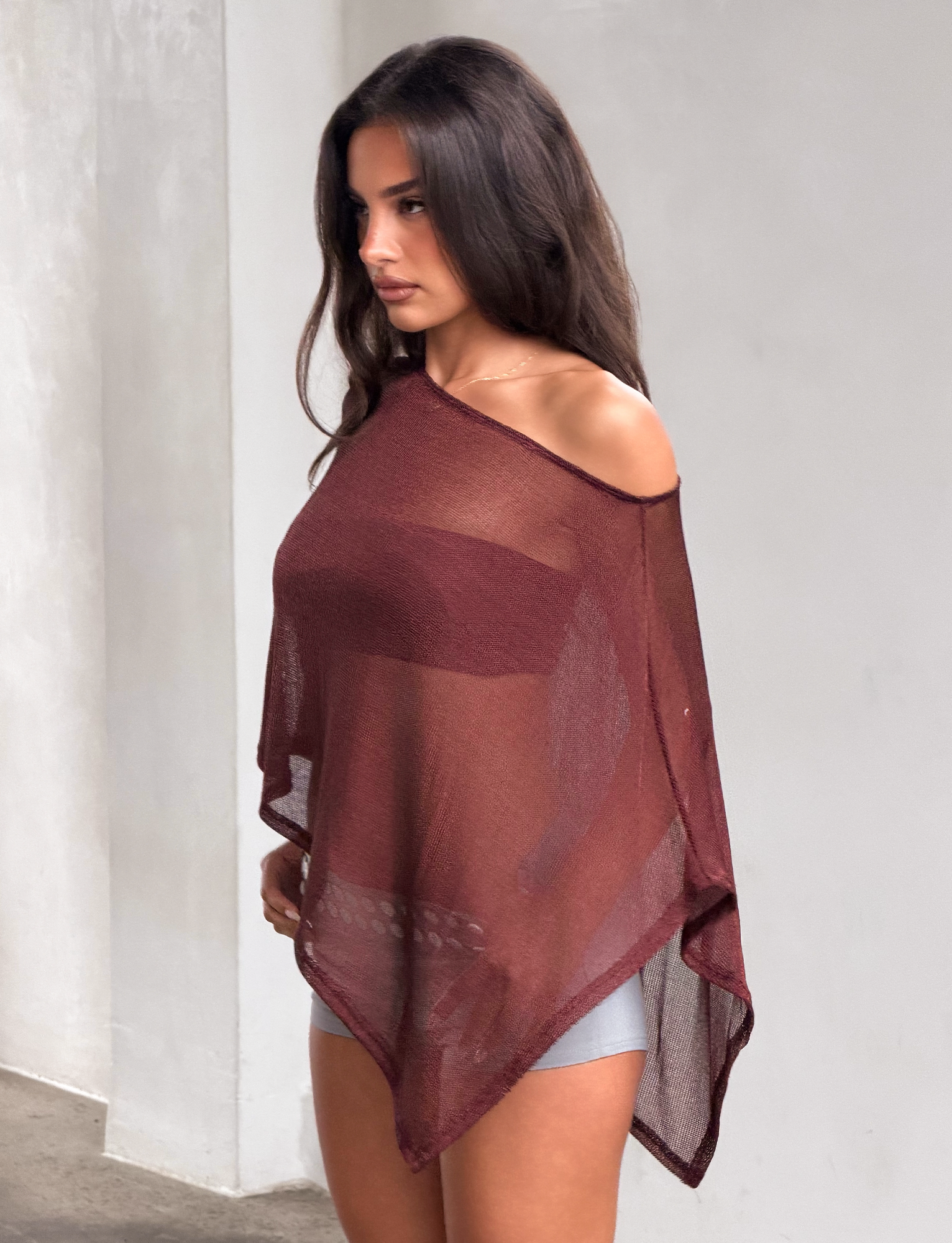 A woman with long brown hair wears the TIGERMIST RAFAELA PONCHO TOP in chocolate over light shorts, standing against a plain, light-colored wall and looking to her left.