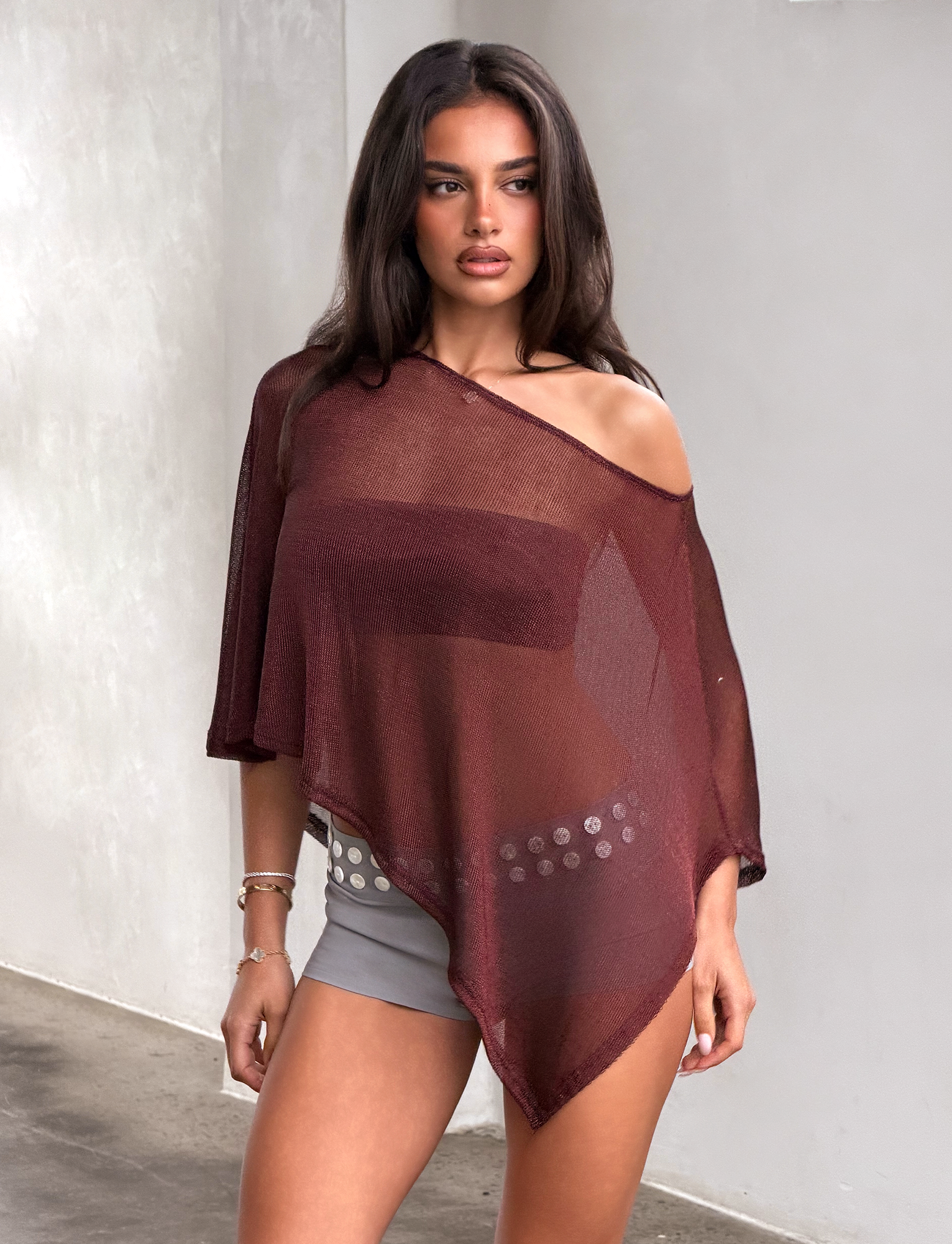A woman stands indoors wearing the TIGERMIST RAFAELA PONCHO TOP in chocolate, layered over a strapless bra and paired with gray shorts featuring a silver belt, looking to the side with a neutral expression.