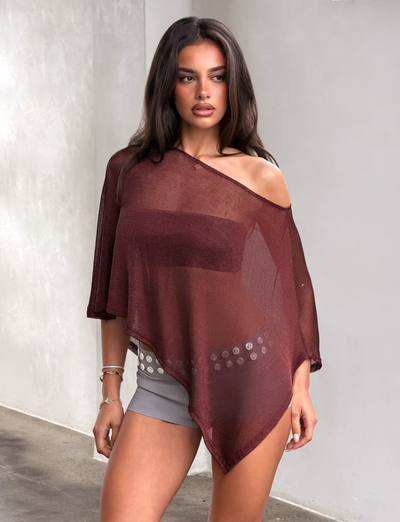 A woman stands indoors wearing the TIGERMIST RAFAELA PONCHO TOP in chocolate, layered over a strapless bra and paired with gray shorts featuring a silver belt, looking to the side with a neutral expression.