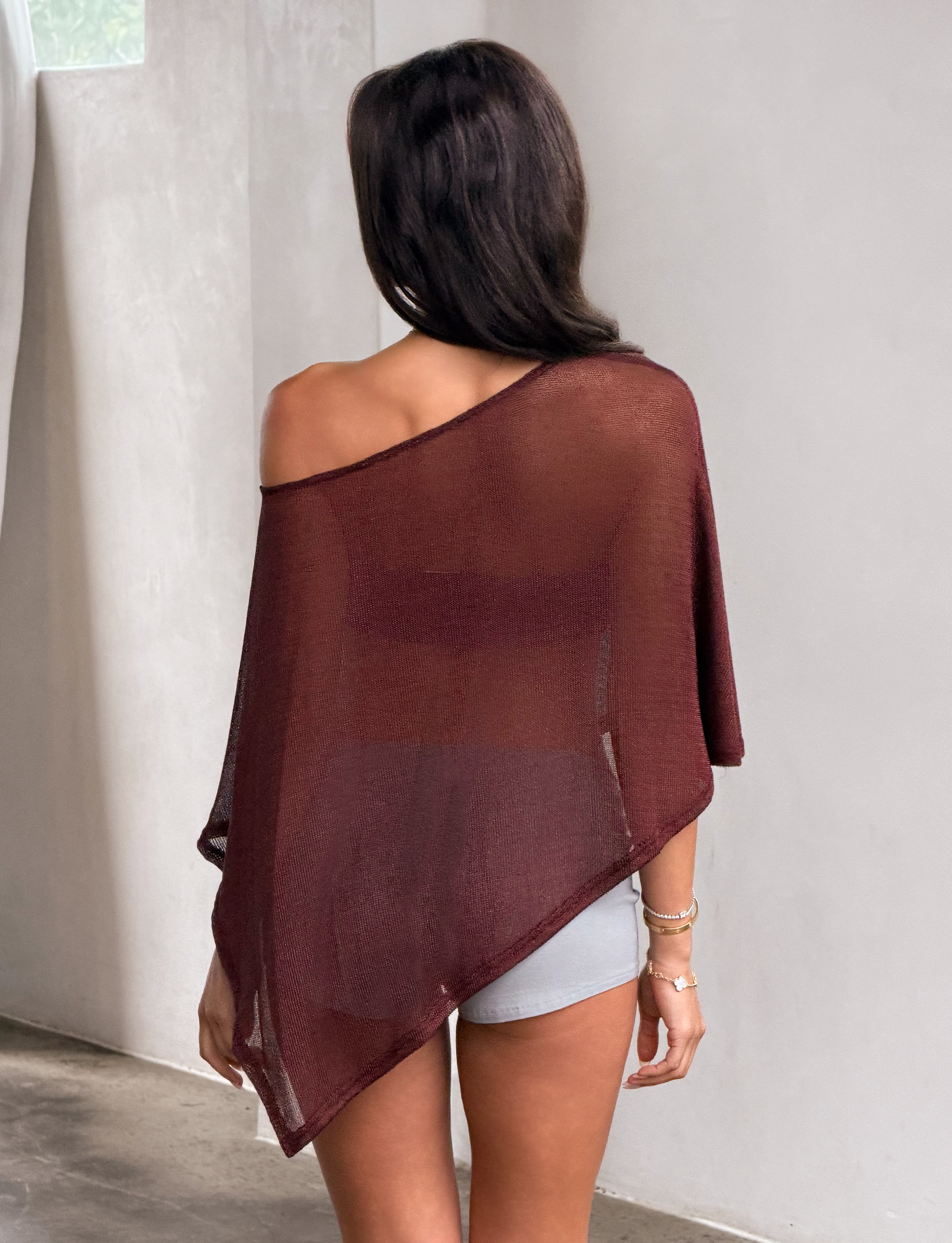 A woman stands indoors facing away, wearing the RAFAELA PONCHO TOP in chocolate by TIGERMIST over light shorts. Her right shoulder is exposed and sunlight filters in from a window on the left.
