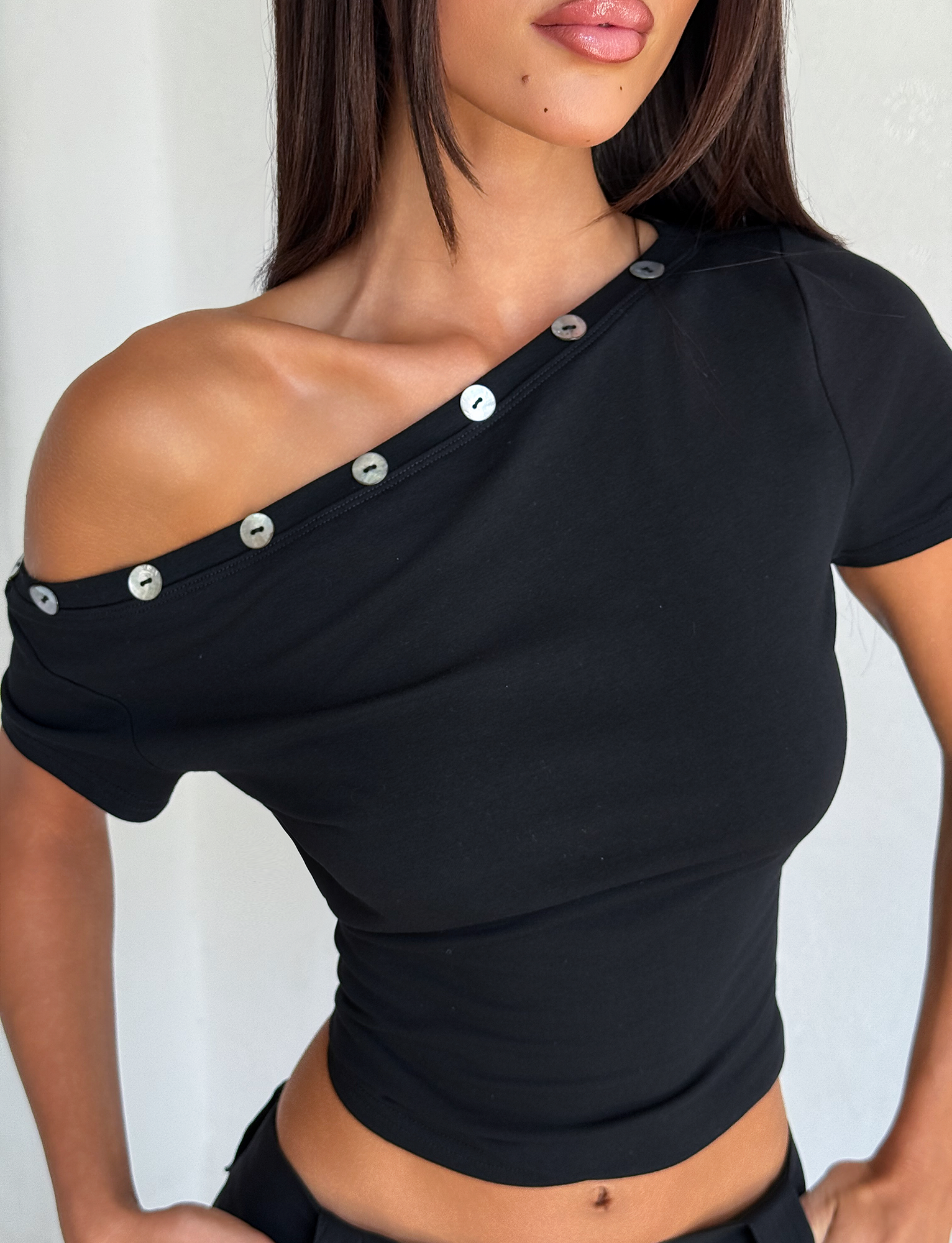 A woman wears the TIGERMIST MARITA TOP in black, a fitted off-the-shoulder tee with short sleeves and shell buttons along the neckline, exposing one shoulder against a plain light background.