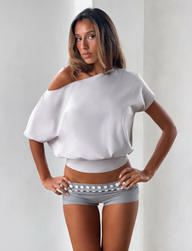 A woman with long brown hair stands with hands on hips against a plain background, wearing the TIGERMIST DRELLA TOP in dove grey—a loose, blouson silhouette—paired with tight gray shorts featuring a white decorative band.