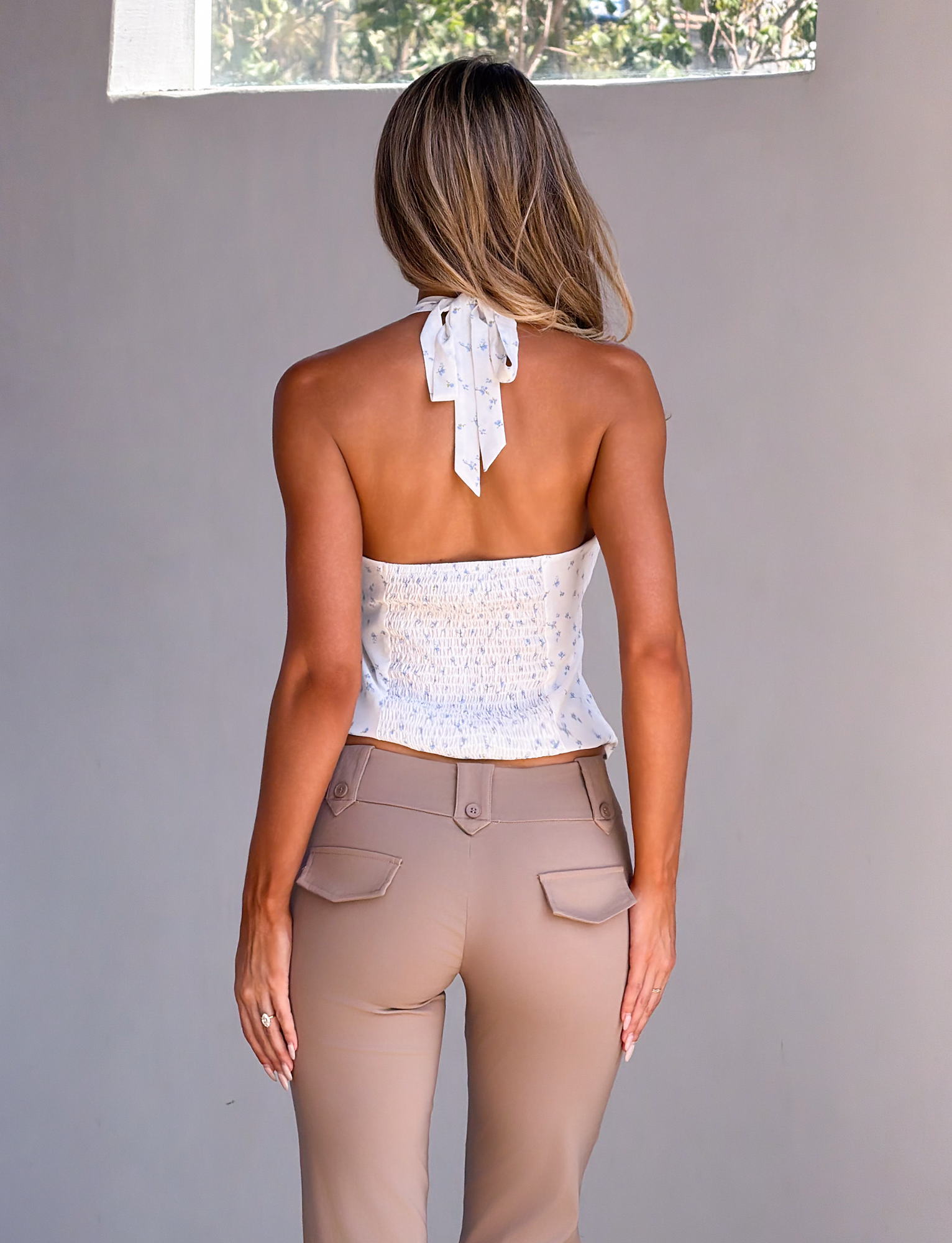 A woman with long, wavy hair faces away, wearing the TIGERMIST CALI TOP - DITSY FLORAL and beige high-waisted pants, standing in front of a plain light wall with a window above.