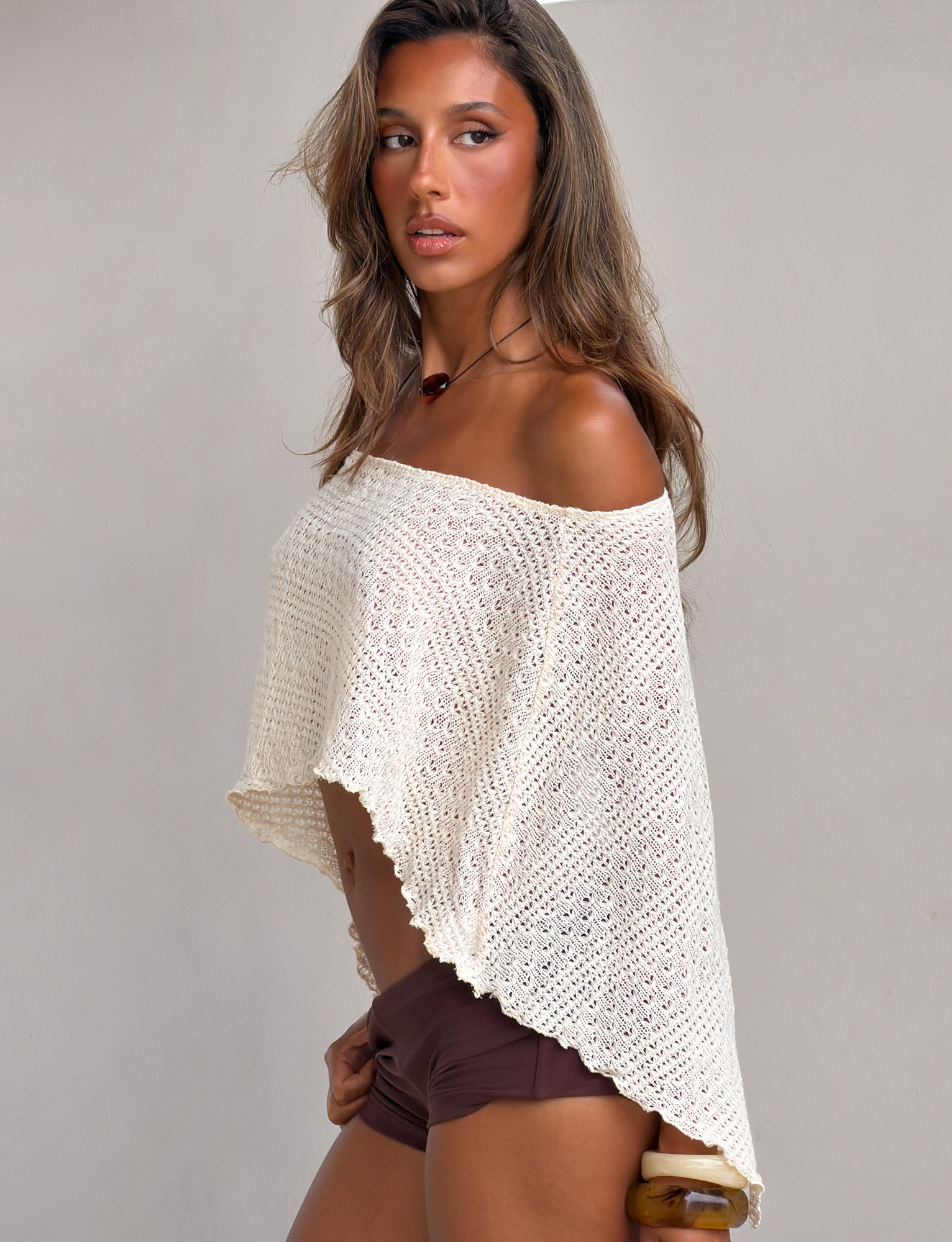 A woman with long brown hair wears the TIGERMIST FAIRLEE PONCHO in cream and brown shorts, standing against a neutral background and gazing to the side.