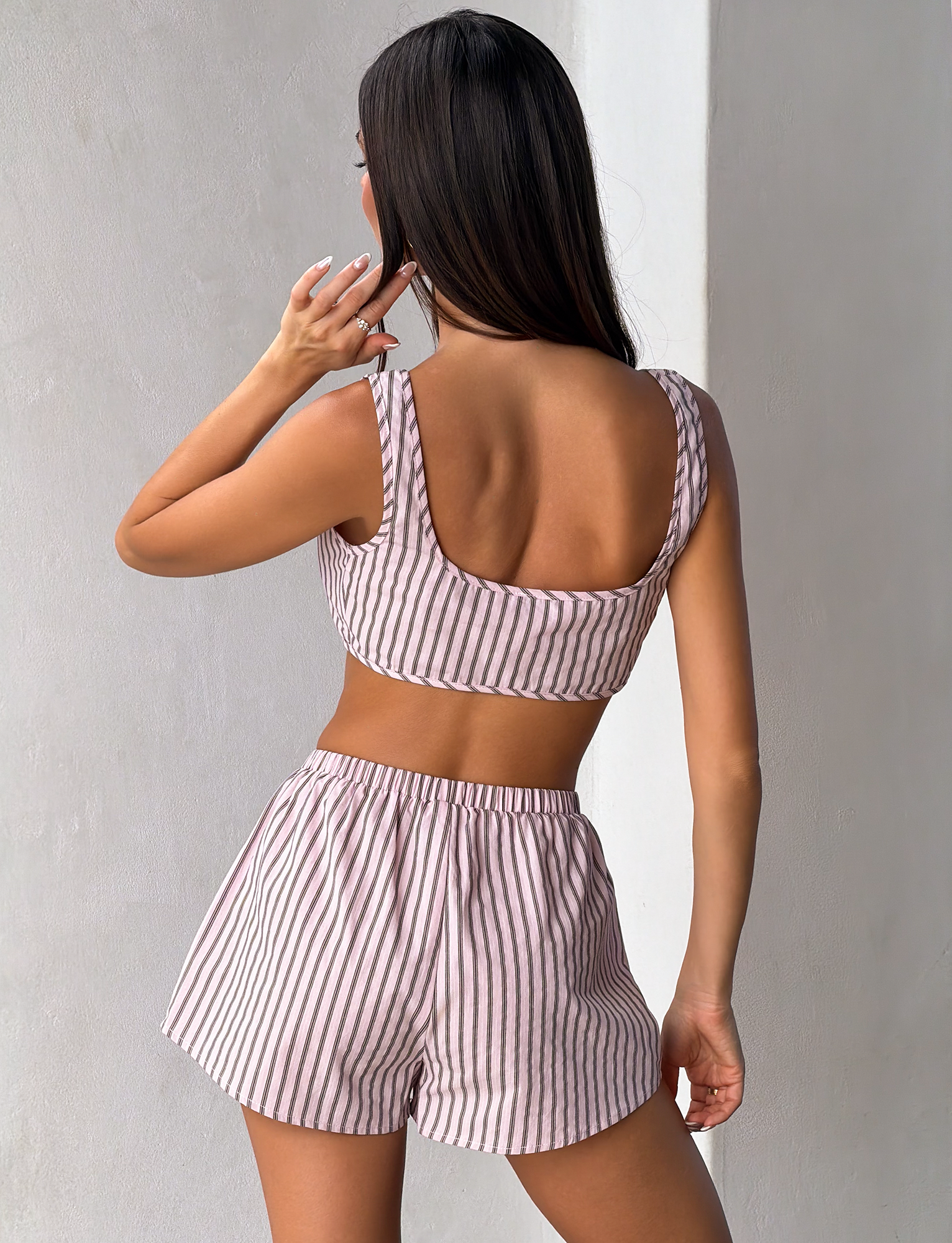 A woman with long brown hair faces a light wall, wearing TIGERMIST’s EZRA SHORT - PINK STRIPE set: a sleeveless striped crop top and matching high-waisted pink and white pinstripe shorts, one hand raised near her face.