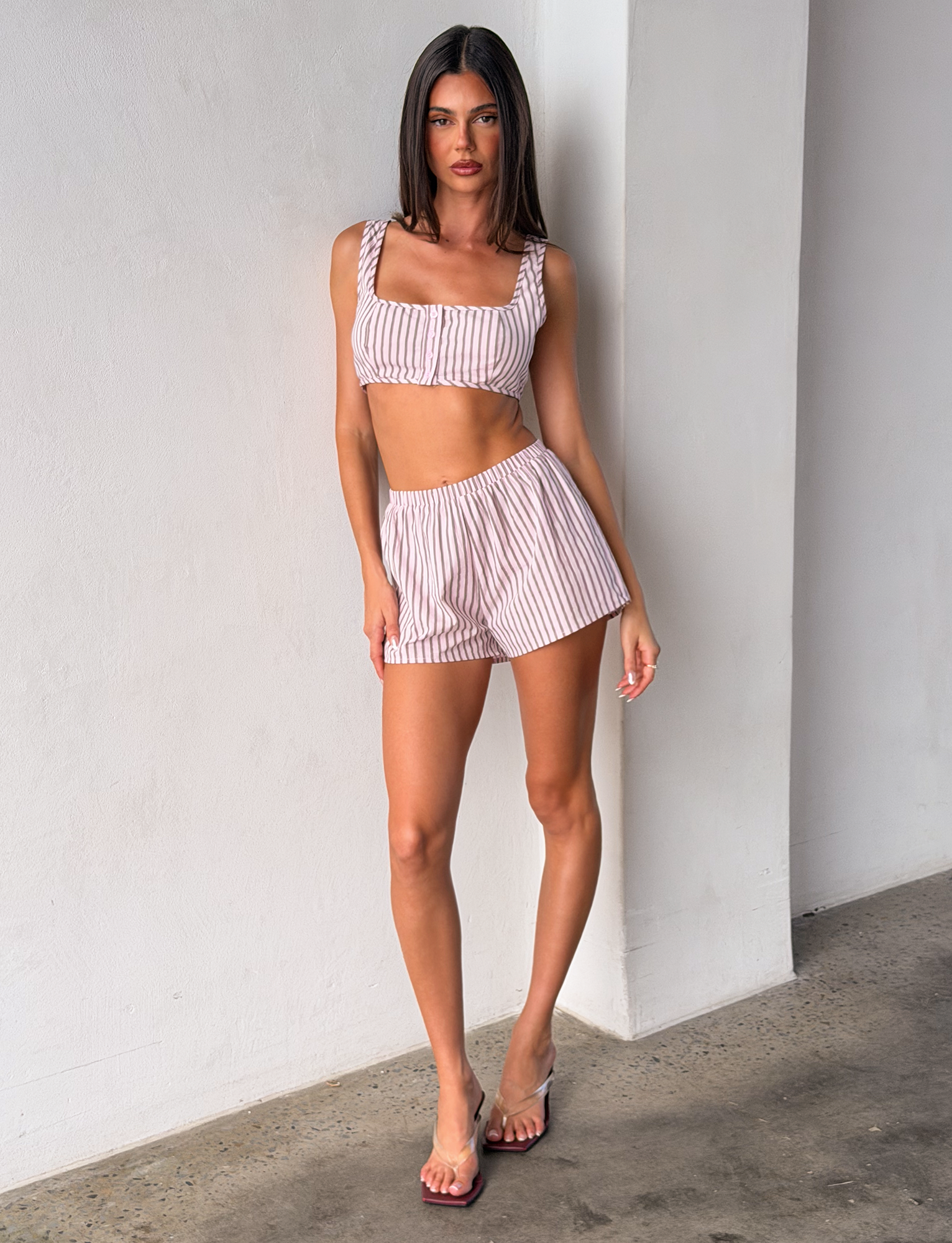 A woman with straight dark hair stands indoors against a white wall, wearing the TIGERMIST EZRA TOP - PINK STRIPE and matching shorts, paired with clear heeled sandals. She has a neutral expression and the floor is concrete.