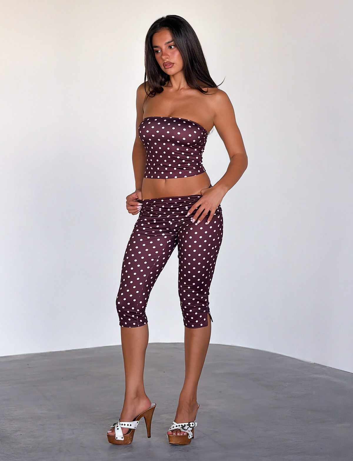 A woman with long dark hair and a neutral expression models the TIGERMIST VALI POLKA DOT TOP in Chocolate Pink Dot, paired with matching bottoms and white high-heeled sandals, standing indoors against a plain light background.