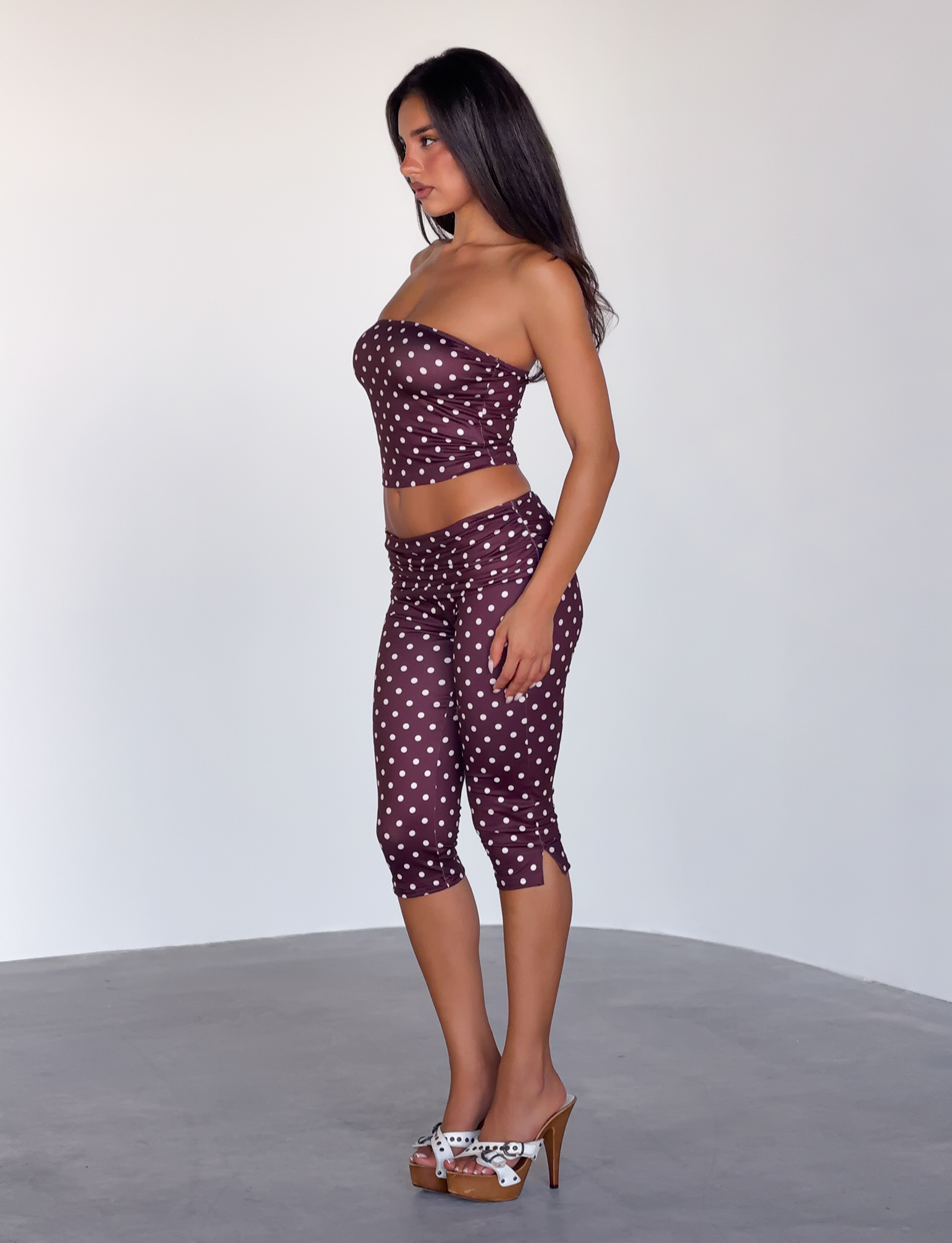 A woman stands in profile against a plain background, wearing the TIGERMIST VALI POLKA DOT CAPRI in chocolate pink dot, paired with a maroon strapless top and high-heeled sandals featuring a white pattern.