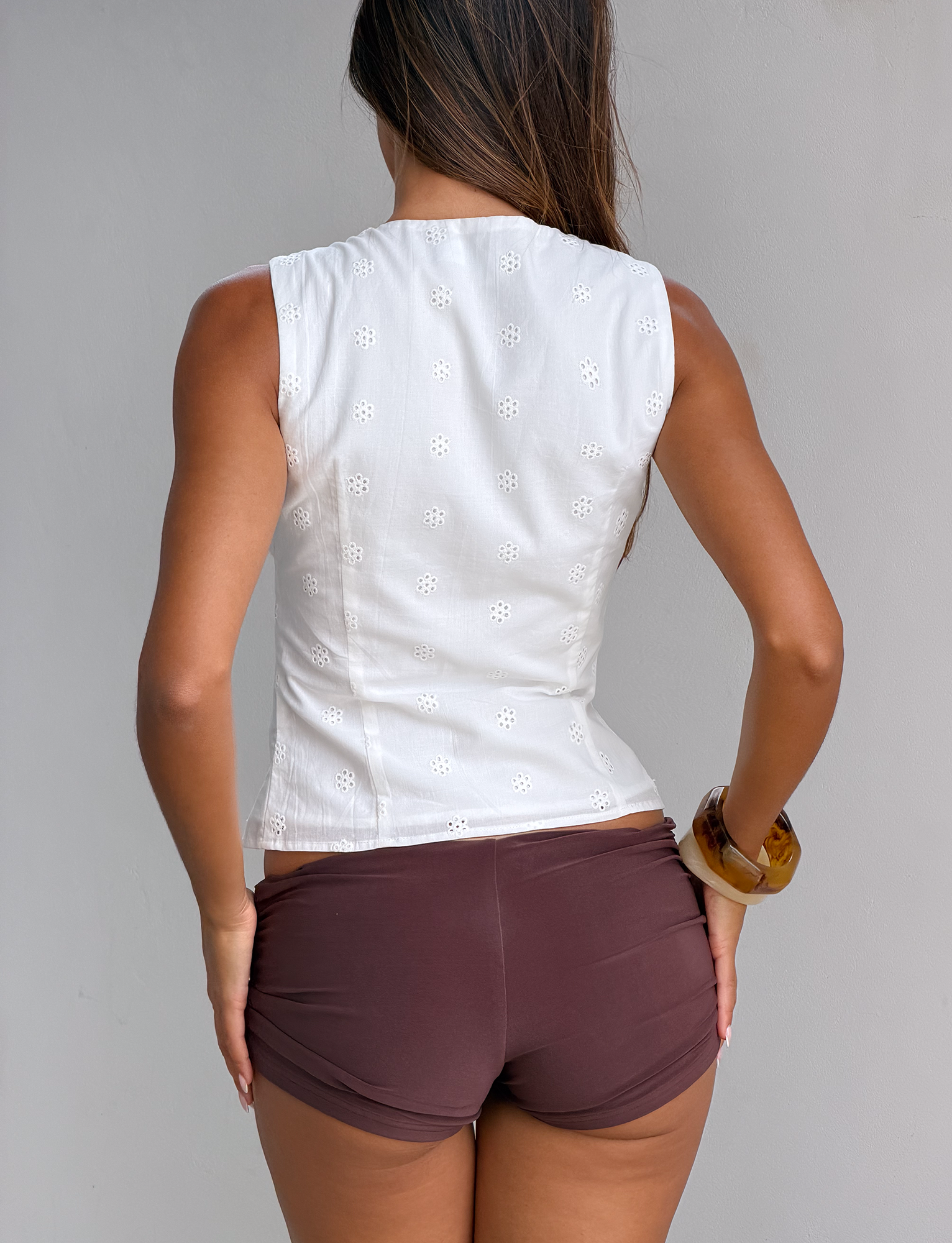 A woman with long brown hair faces away, wearing the TIGERMIST CAELYNN TOP in white with eyelet trim and brown shorts. She accessorizes with a large bracelet, standing before a neutral-colored wall.