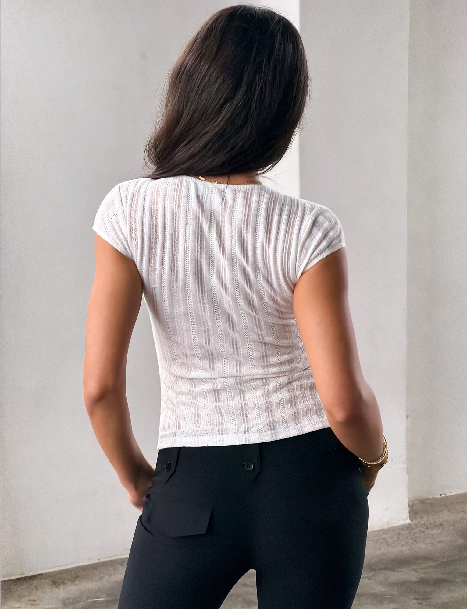 A woman with long brown hair stands with her back to the camera, wearing the TIGERMIST LONA TOP in white and black pants, indoors against a plain light-colored wall.