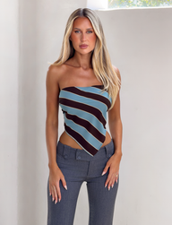 A woman with long blonde hair wears the TIGERMIST LENIN SCARF TOP - BLUE AND BROWN STRIPE, styled strapless with high-waisted gray pants, standing indoors against a light wall and looking directly at the camera.
