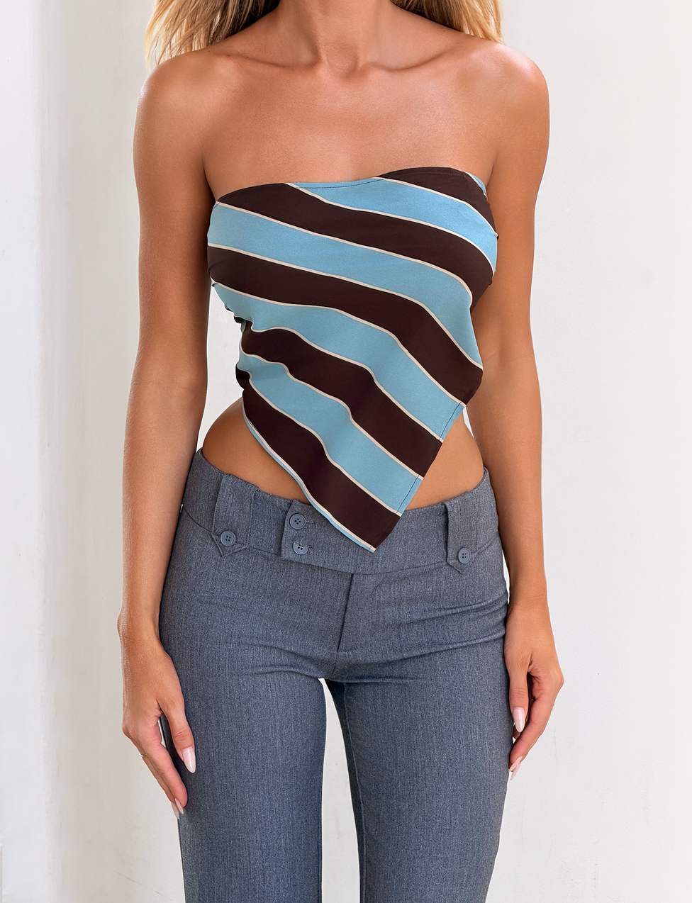 A woman models the TIGERMIST LENIN SCARF TOP - BLUE AND BROWN STRIPE, paired with gray pants, against a plain white background. Her head is not visible in the image.