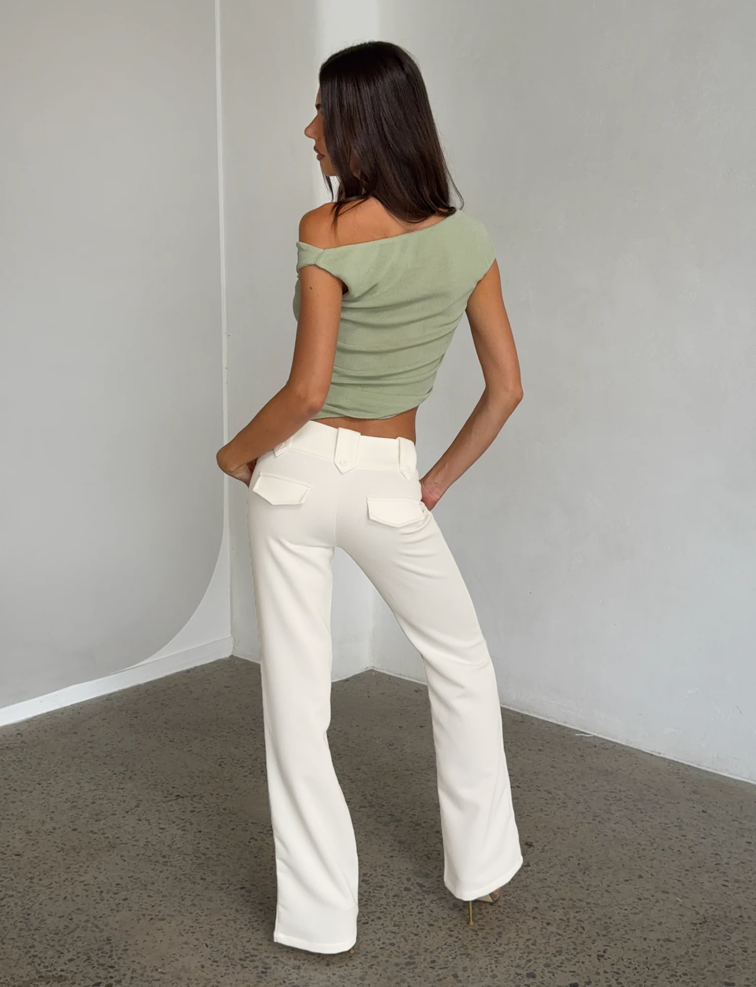 A woman stands indoors on a speckled floor, facing away. She wears a light green off-shoulder crop top and the TIGERMIST KITTIE PANT - WHITE : CREAM in a high-waisted, flare fit. The background is minimal with light gray walls.