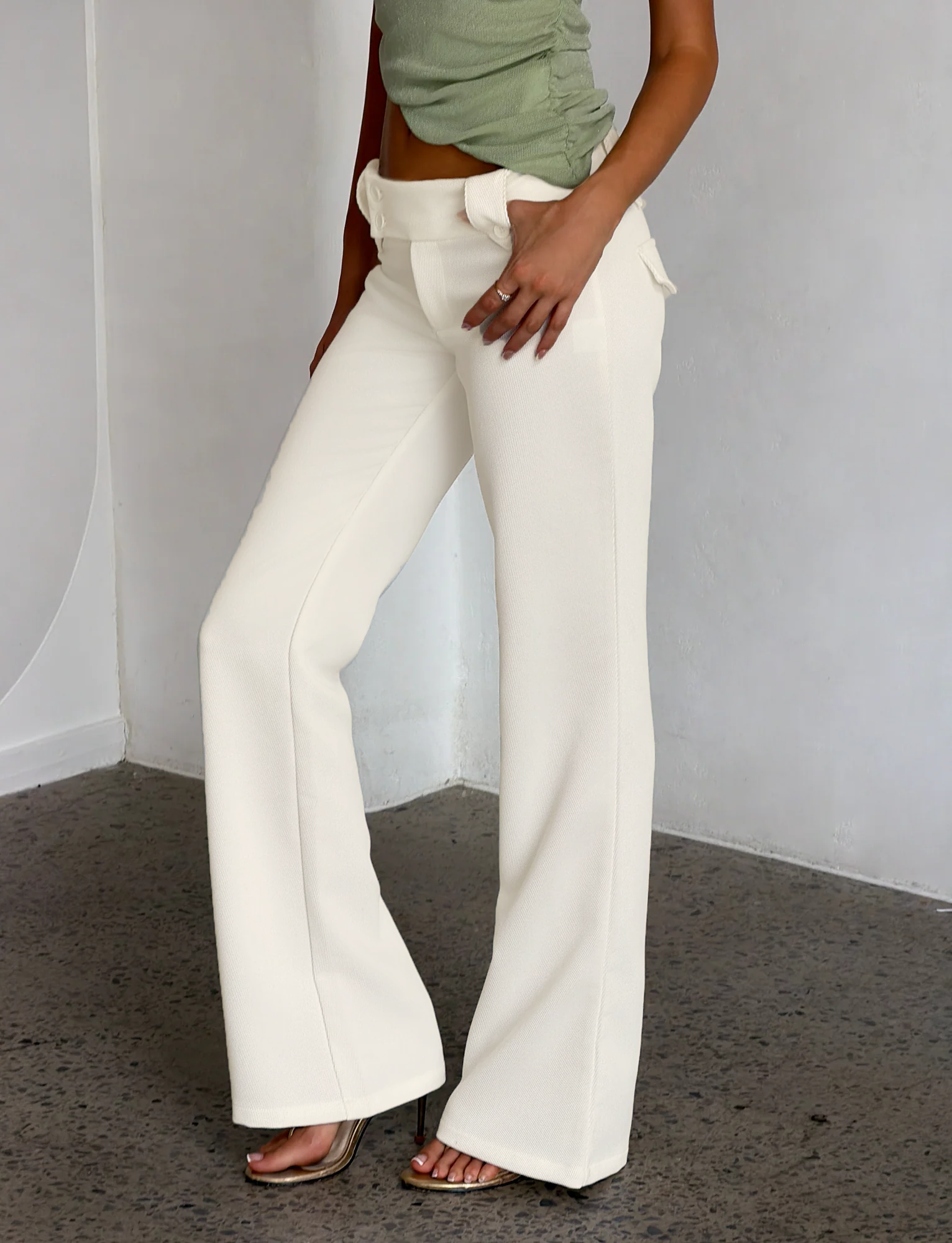 A person stands indoors on a polished concrete floor, wearing TIGERMIST's KITTIE PANT - WHITE : CREAM high-waisted flare pants with a light green sleeveless top, and rests one hand in a pocket.