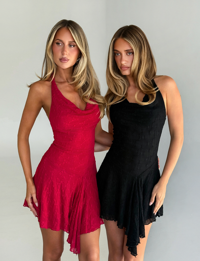 Two women with long blonde hair wear asymmetrical dresses from TIGERMIST, including the FIORELLA DRESS - RED LACE, standing side by side against a plain light background and looking slightly away from the camera.