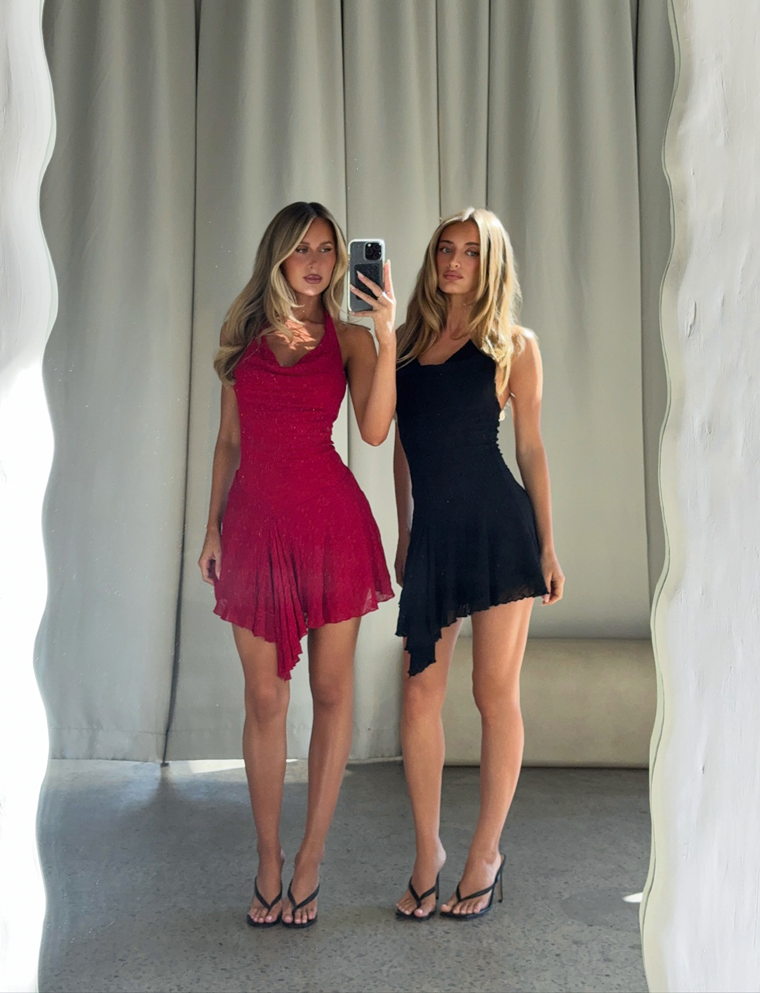Two women with long blonde hair pose for a mirror selfie. One wears the TIGERMIST FIORELLA DRESS - RED LACE, the other a black dress. Both wear black sandals. Light curtains hang in the background.