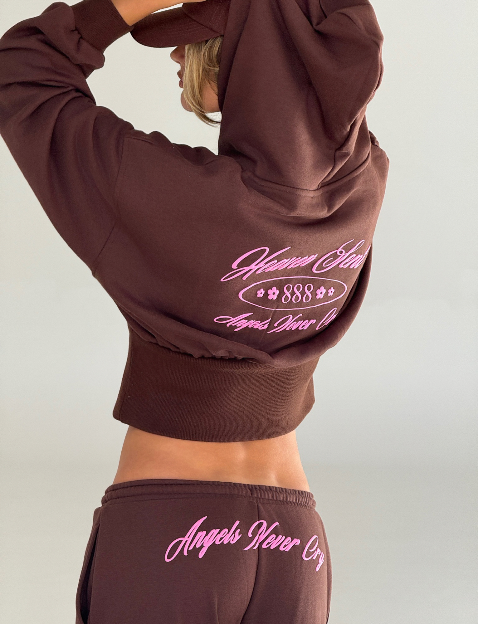 A person in the TIGERMIST MERRYN HOODIE - CHOCOLATE and matching pants with pink "Heaven Sent 888 Angels Never Cry" embroidery on the hoodie and "Angels Never Cry" on the pants, standing sideways adjusting the hood.