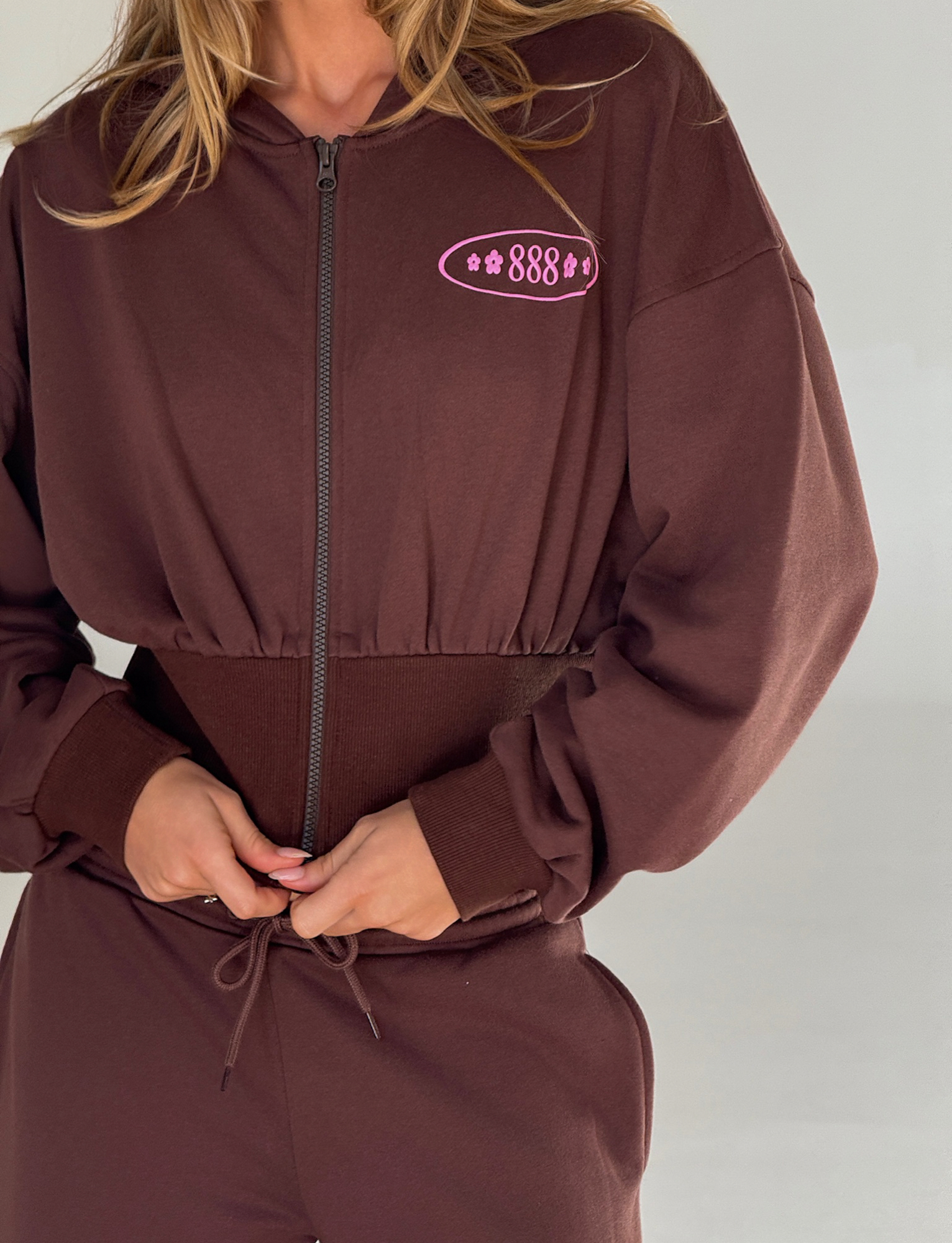 A person in the TIGERMIST MERRYN HOODIE - CHOCOLATE and matching pants adjusts the waistband. The hoodie has a pink "888" puff print with stars on the chest, set against a plain, light background.