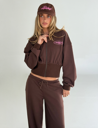 A woman with long blonde hair stands against a light grey background wearing the TIGERMIST MERRYN HOODIE in chocolate, paired with matching sweatpants and a brown baseball cap, resting one hand near her chin.