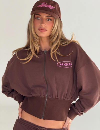 A woman with long blonde hair models the TIGERMIST MERRYN HOODIE in chocolate, featuring pink puff print details, with a matching cap. She poses with her hand on her hip against a plain light background.