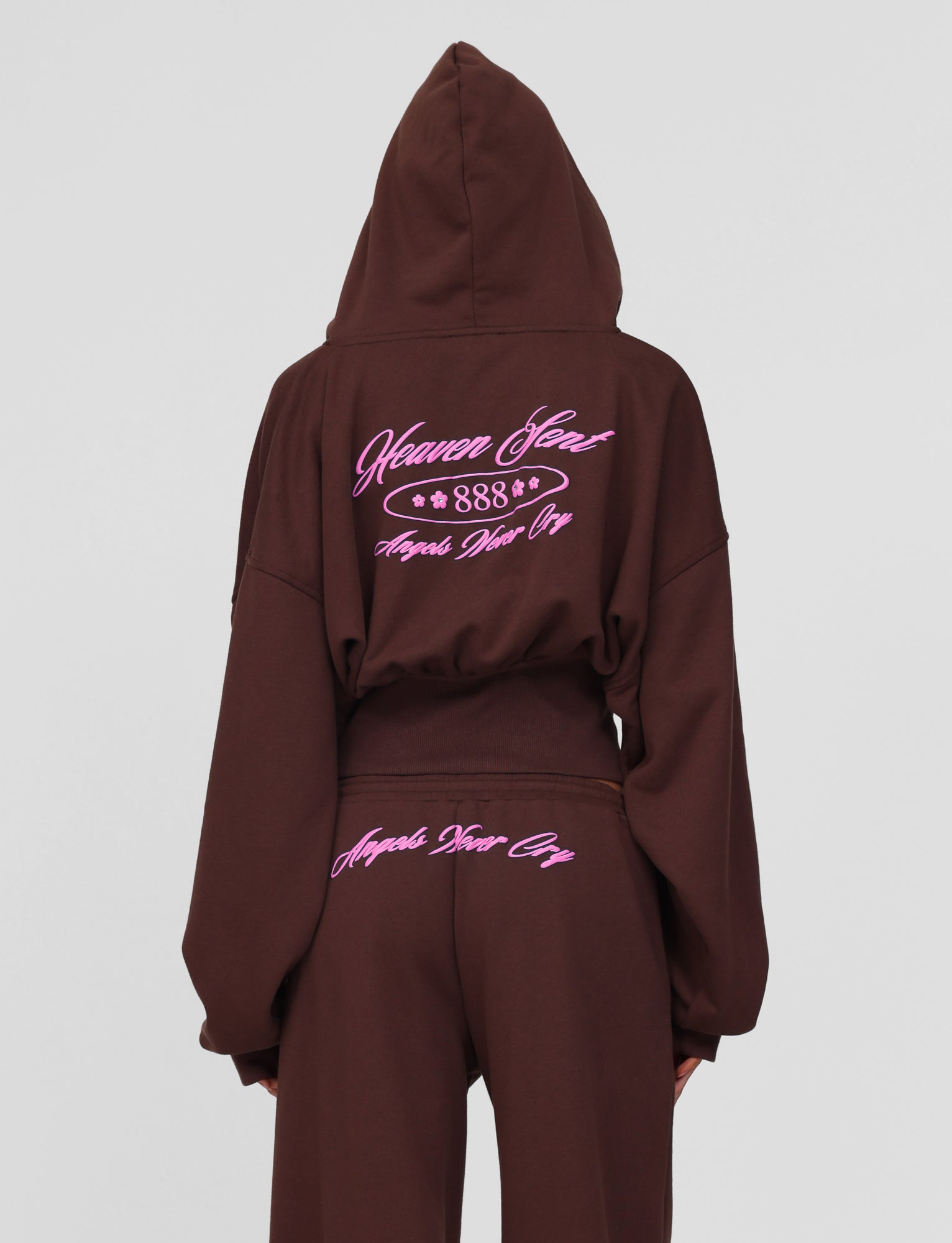 A person faces away from the camera wearing the TIGERMIST MERRYN HOODIE in chocolate and matching pants, both with pink embroidered text: "Heaven Sent 888 Angels Work Overtime" on the hoodie and "Angels Never Cry" on the pants.