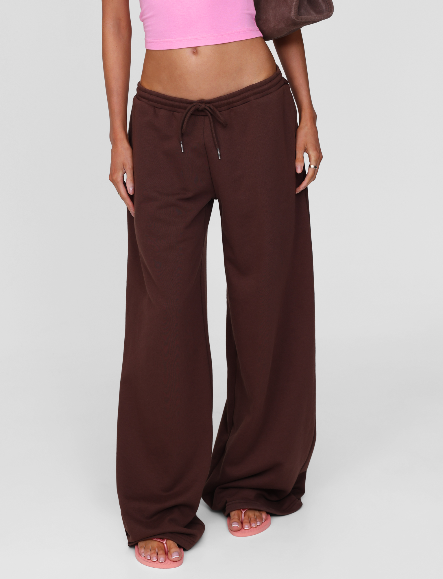 A person wears TIGERMIST's MERRYN TRACK PANT in chocolate and a pink crop top, standing barefoot with a brown bag, cropped at the shoulders, capturing the effortless style of Angels Never Cry.