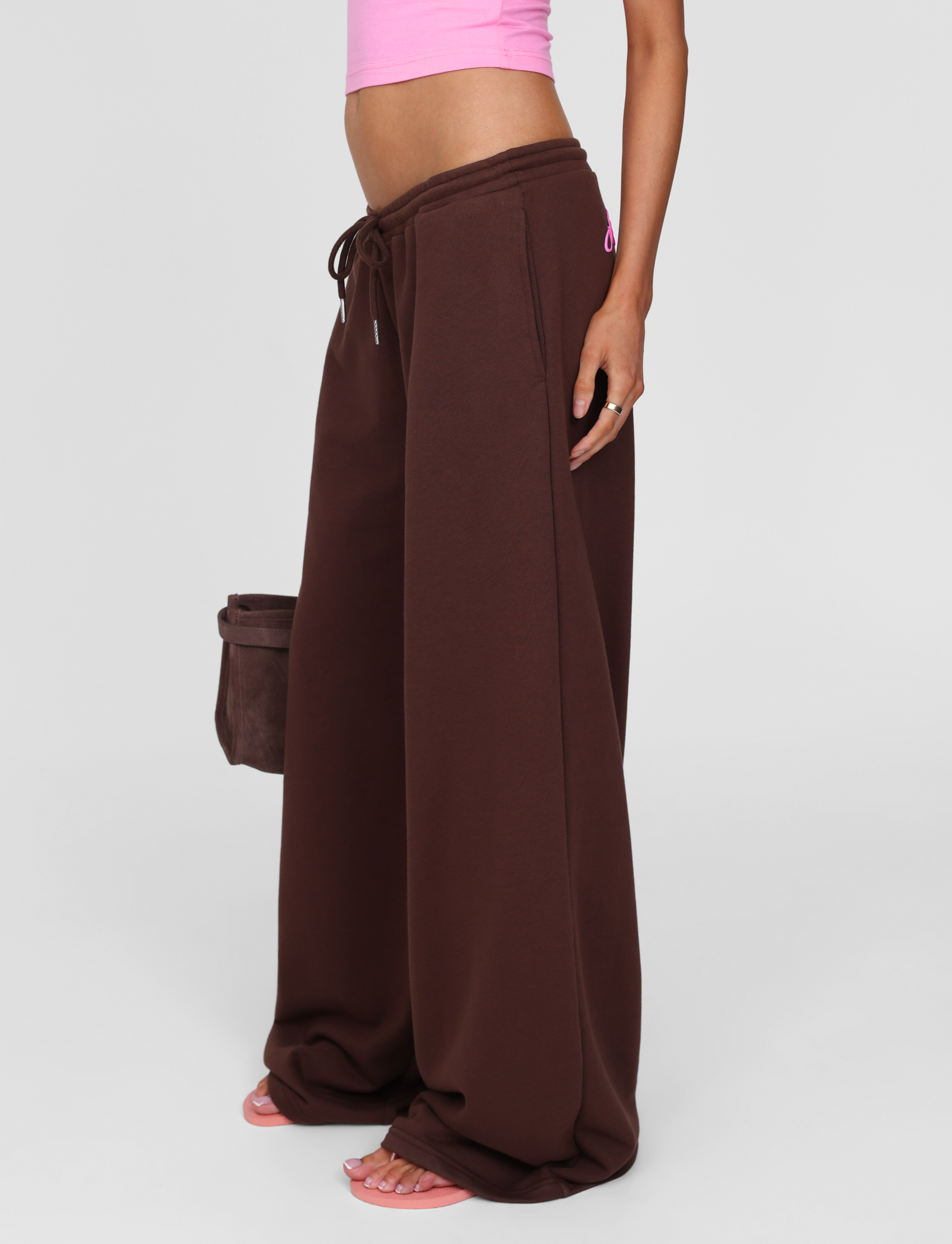 A person styled in TIGERMIST's MERRYN TRACK PANT - CHOCOLATE, paired with a pink crop top, pink sandals, and a brown handbag stands against a light gray backdrop—urban chic inspired by Angels Never Cry.