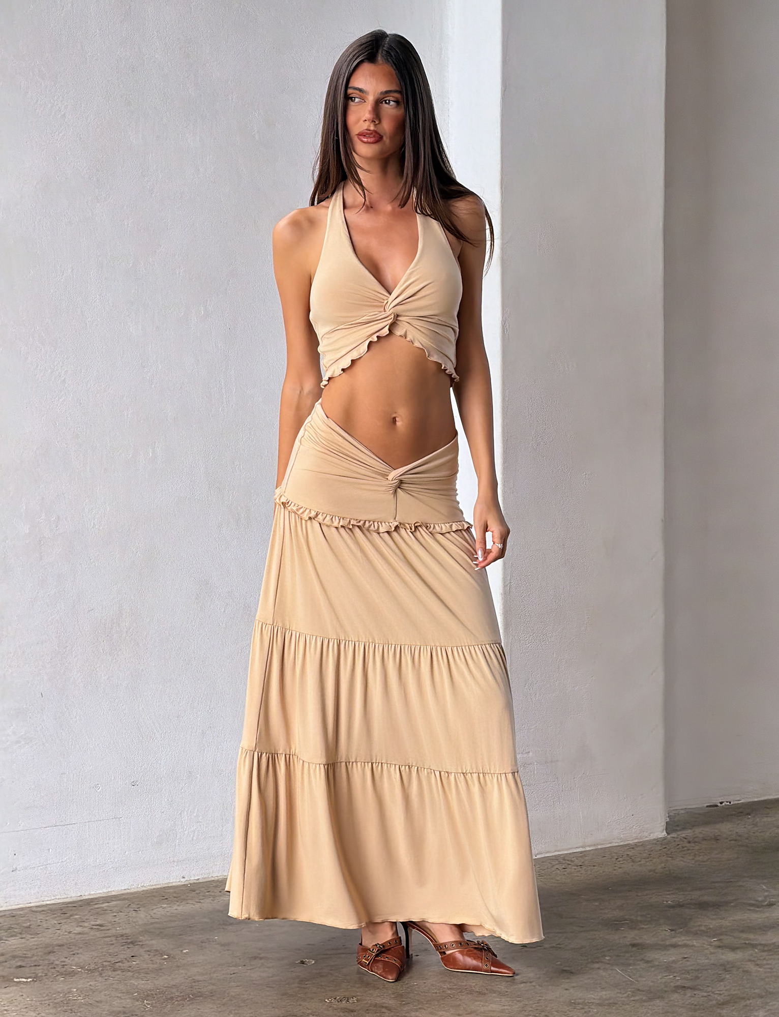 A woman stands indoors wearing the TIGERMIST KATYA MAXI SKIRT in tan with a matching beige halter crop top. She has long straight hair, minimal makeup, and brown flat shoes against a plain light-colored background.