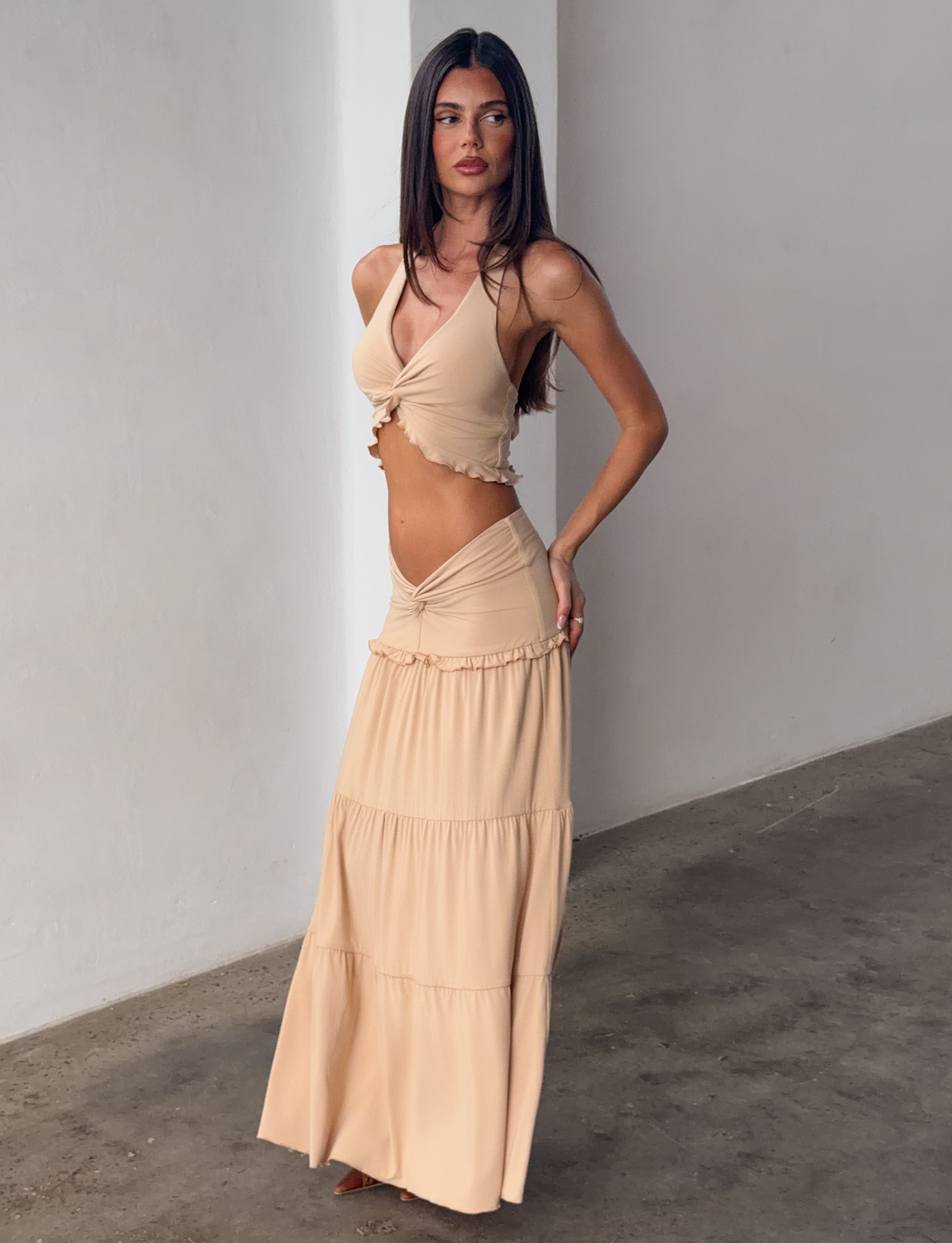 A woman with long dark hair poses indoors against a plain wall, wearing the TIGERMIST KATYA MAXI SKIRT in tan with a matching light beige sleeveless crop top, her hand on her hip as she looks to the side.