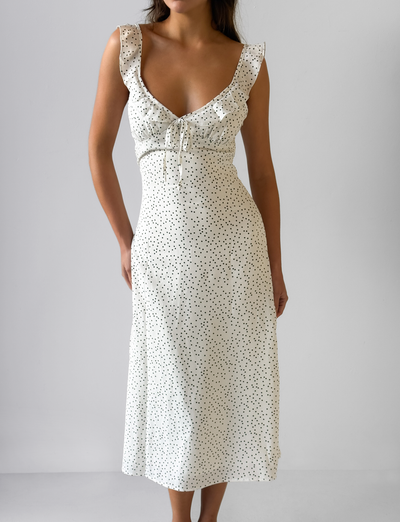 A woman wears the CL - LOTTIE RUFFLE MIDI DRESS by Tiger Mist USA, a sleeveless white midi dress with black polka dots and a ruffled neckline, standing against a plain light gray background.