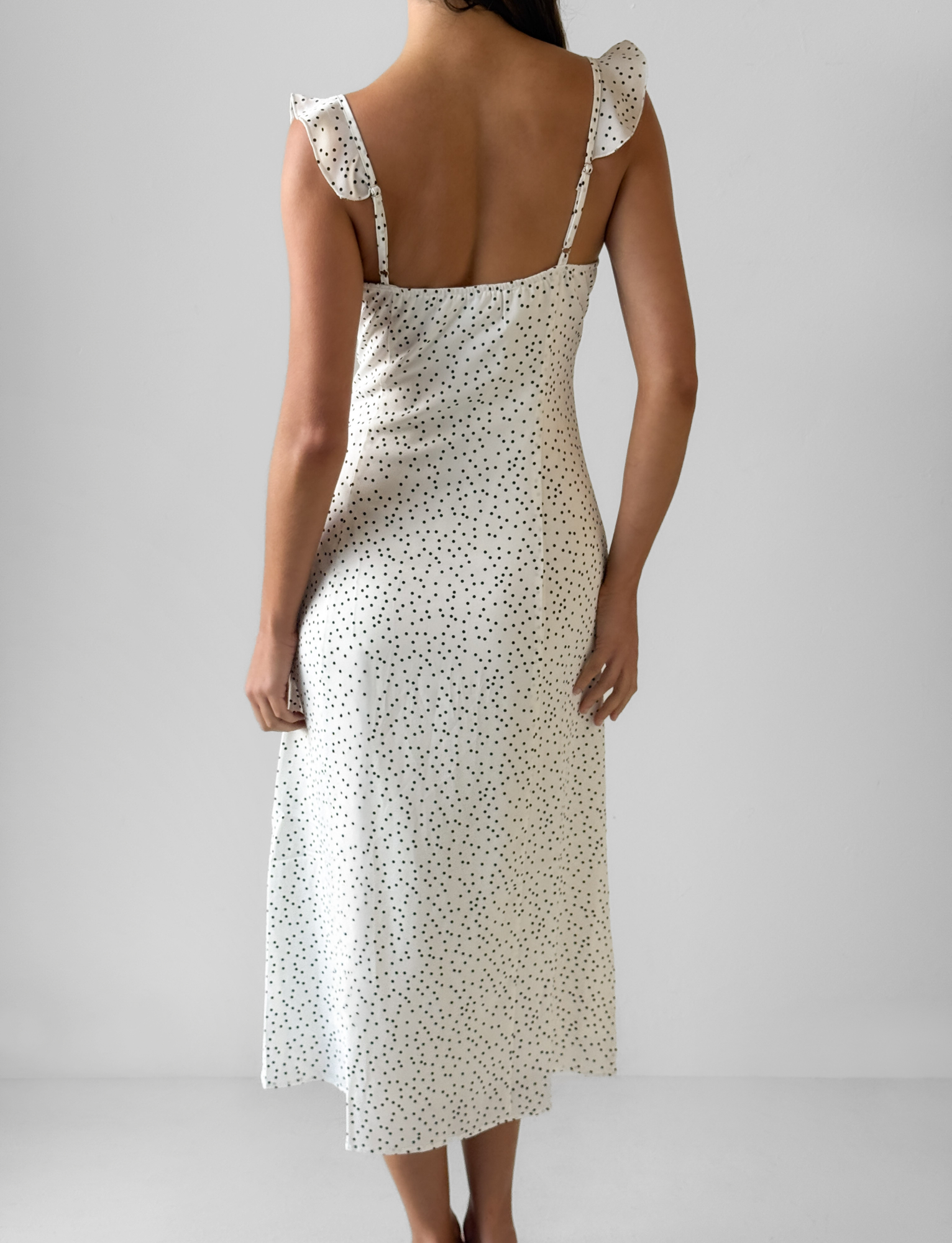 A woman faces away, wearing the Third Party LOTTIE RUFFLE MIDI DRESS - WHITE POLKA with ruffle-trimmed straps, set against a plain light gray background.