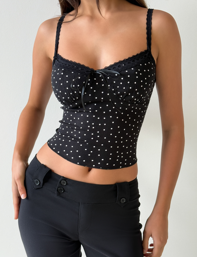 A person wears the Tiger Mist USA CL - BAILEE CAMI TOP, a black fitted camisole with polka dots, lace trim, and bow detail, paired with high-waisted black pants against a plain light background.