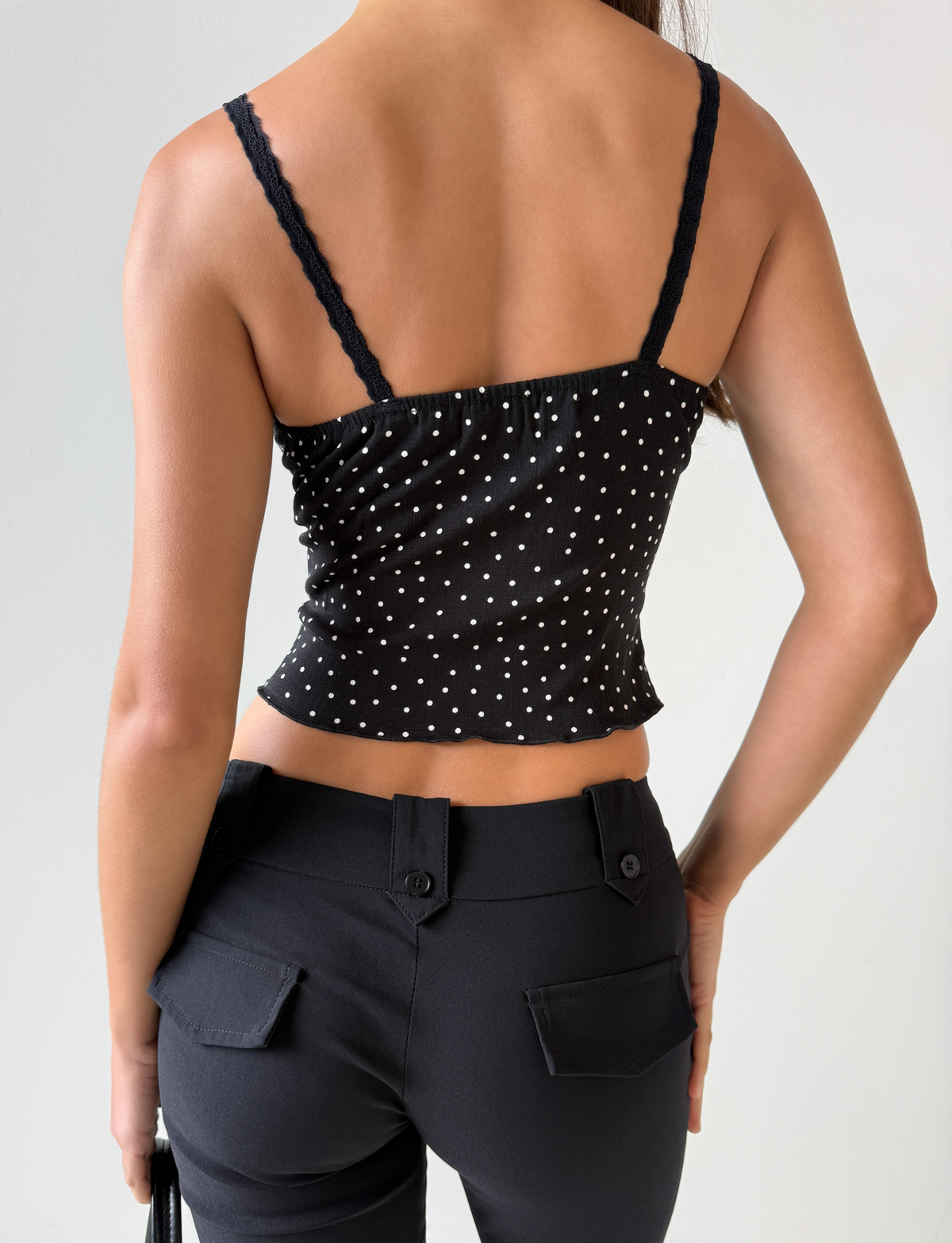 A person is shown from the back wearing the Third Party BAILEE CAMI TOP - BLACK POLKA, featuring white polka dots and lace trim, paired with high-waisted black pants. Their right arm is by their side, and their left hand rests on their hip.