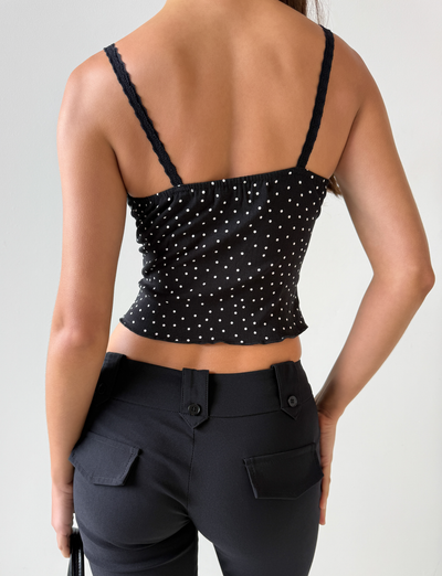 A person is shown from the back wearing the Third Party BAILEE CAMI TOP - BLACK POLKA, featuring white polka dots and lace trim, paired with high-waisted black pants. Their right arm is by their side, and their left hand rests on their hip.